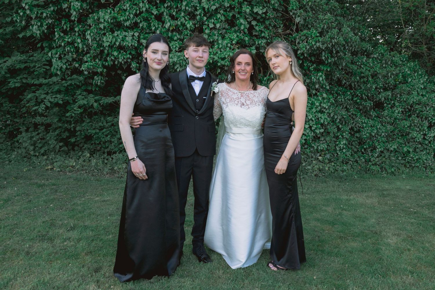 Four teens in formal attire pose outdoors, wearing black gowns and a tuxedo, with lush green foliage behind them.