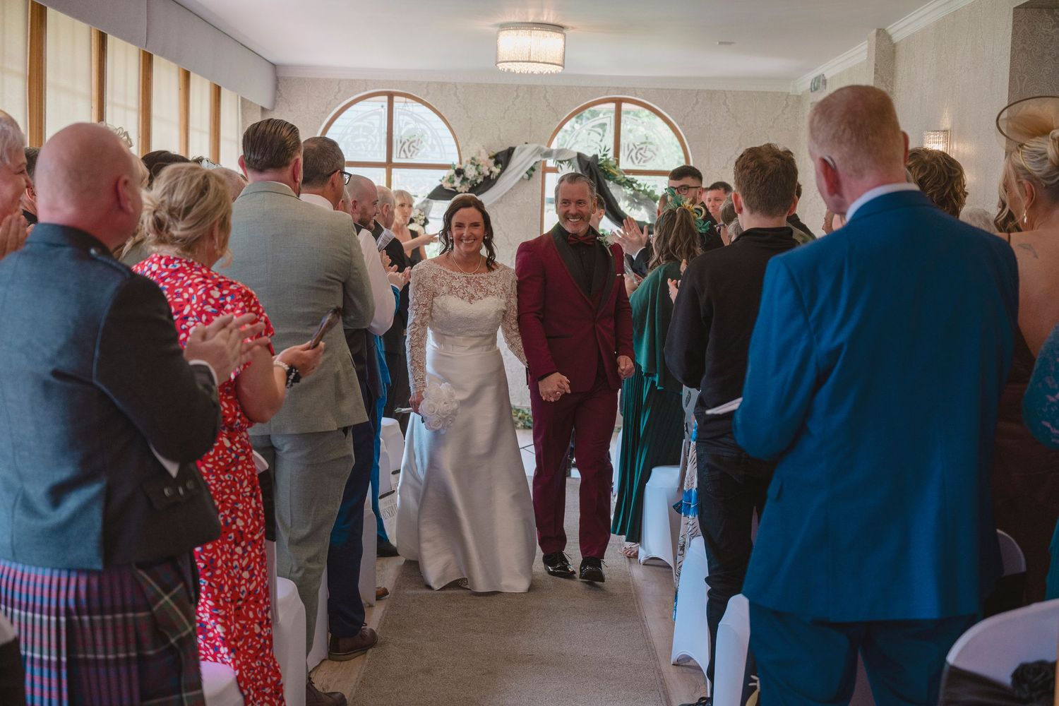 Bride and groom walk down the aisle together, smiling, as guests applaud in a bright wedding venue.
