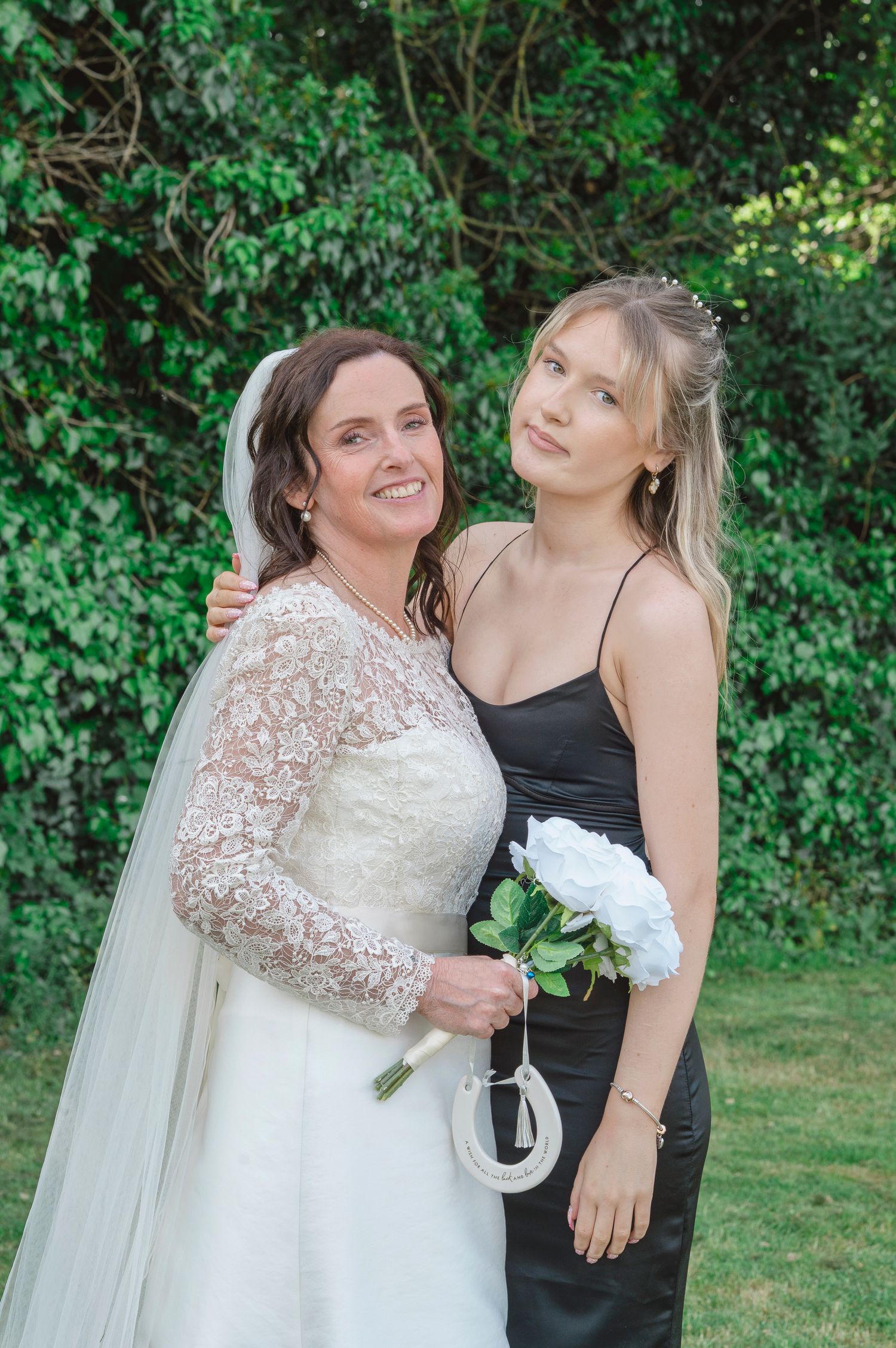 Bride in lace wedding gown and veil poses with bridesmaid in black dress holding white bouquet outdoors.