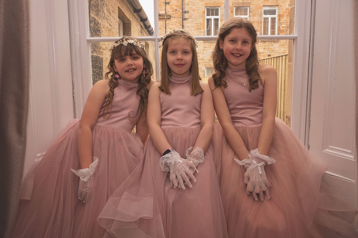 Three young flower girls in matching dusty pink tulle gowns with white gloves and tiaras at a wedding.