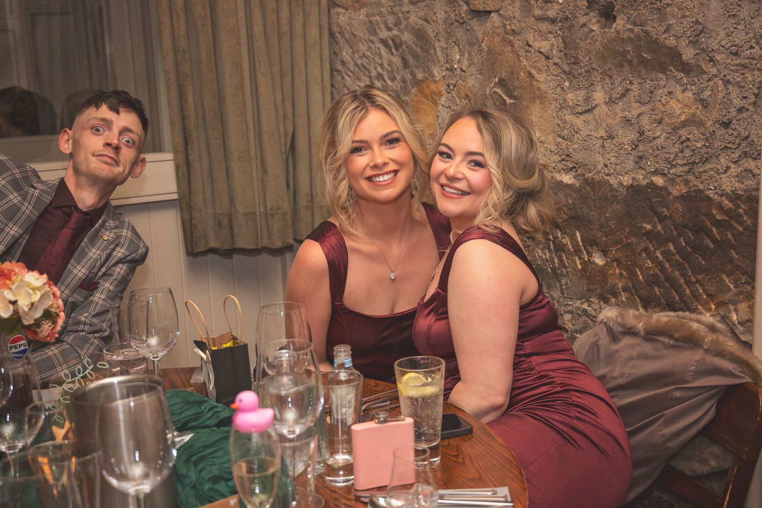Three smiling wedding guests in burgundy and plaid sit together at a reception dinner table.