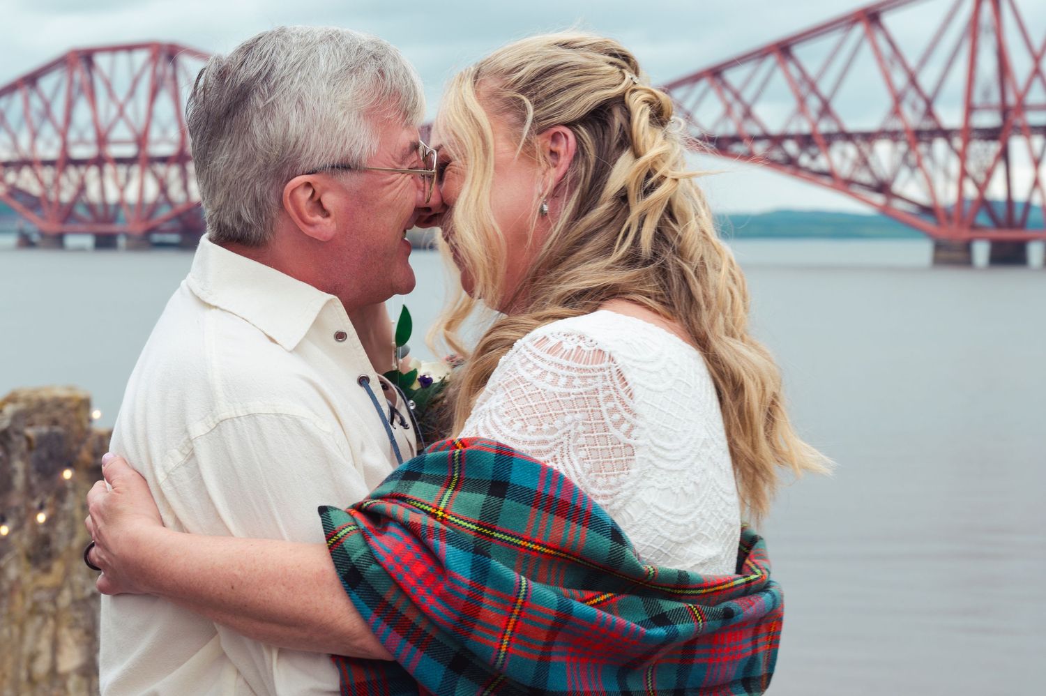 Couple sharing a romantic moment by the Forth Bridge in Scotland, woman draped in colorful tartan plaid.