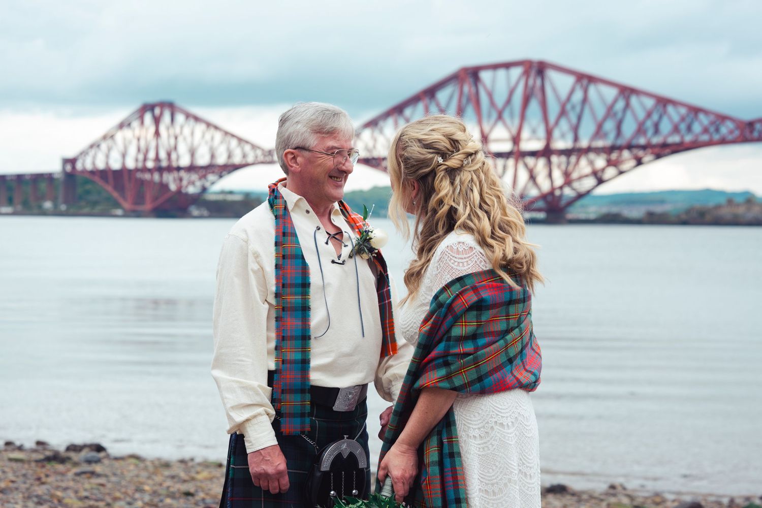 Couple exchanging vows by the Forth Bridge in Scotland, groom in traditional tartan kilt and bride in white lace dress.