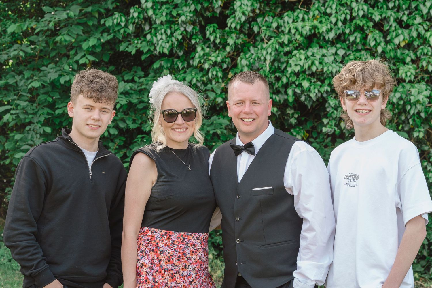 Family of four smiling outdoors, dressed smartly, posing together in front of lush green foliage on a sunny day.