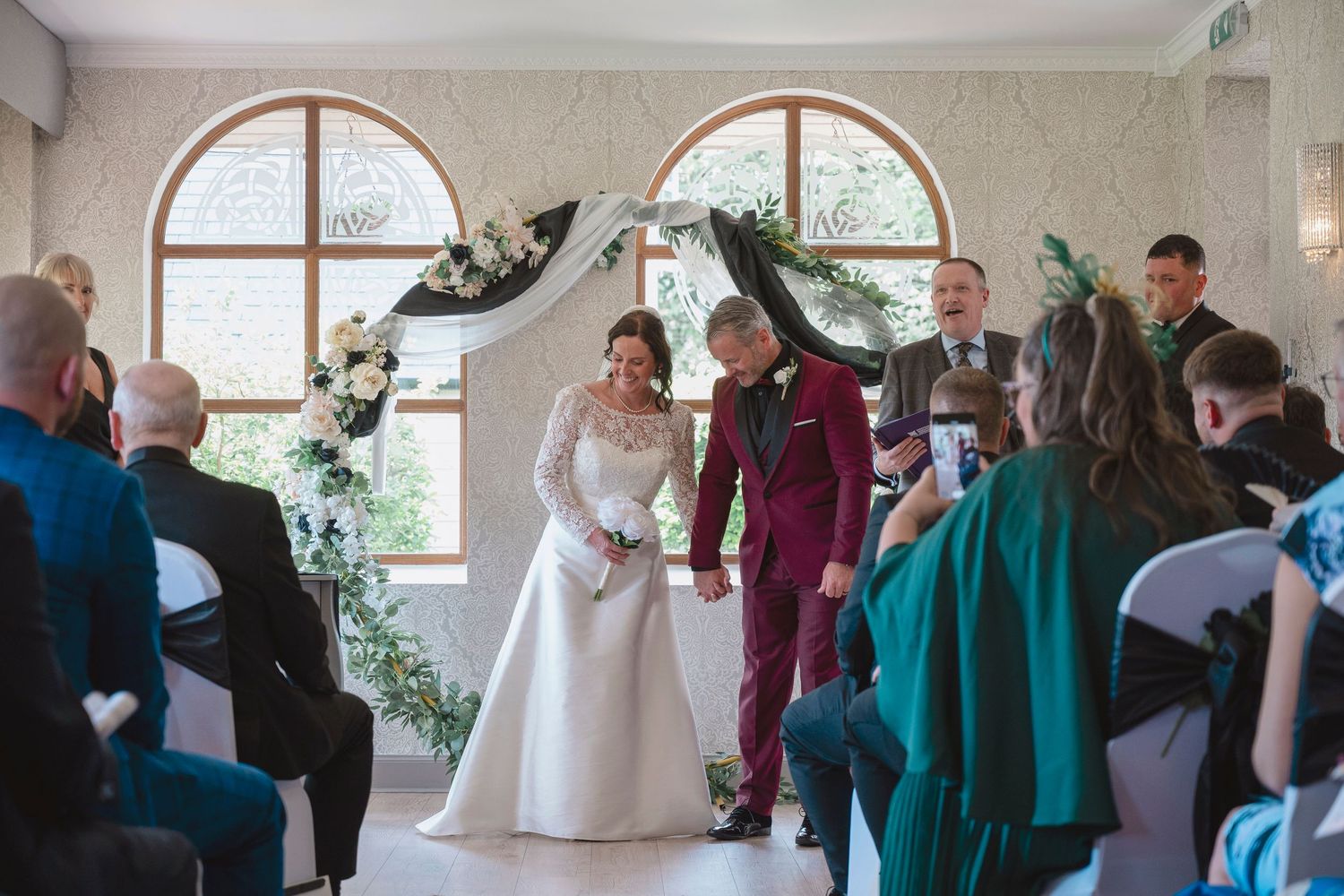 Bride in white gown and groom in burgundy suit hold hands at elegant wedding ceremony with floral arch.