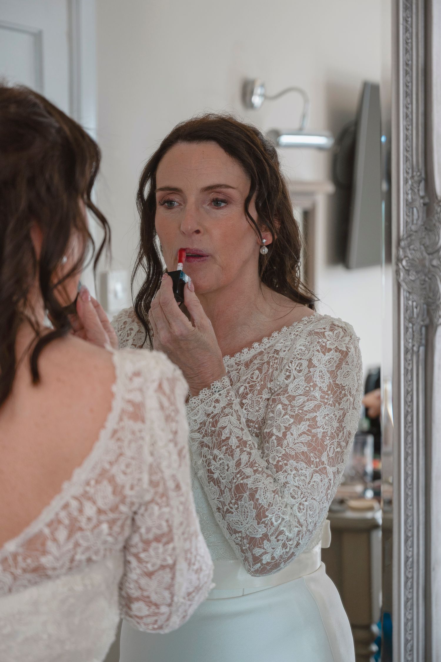 Bride in lace wedding dress applying red lipstick while looking in an ornate mirror during bridal preparations.
