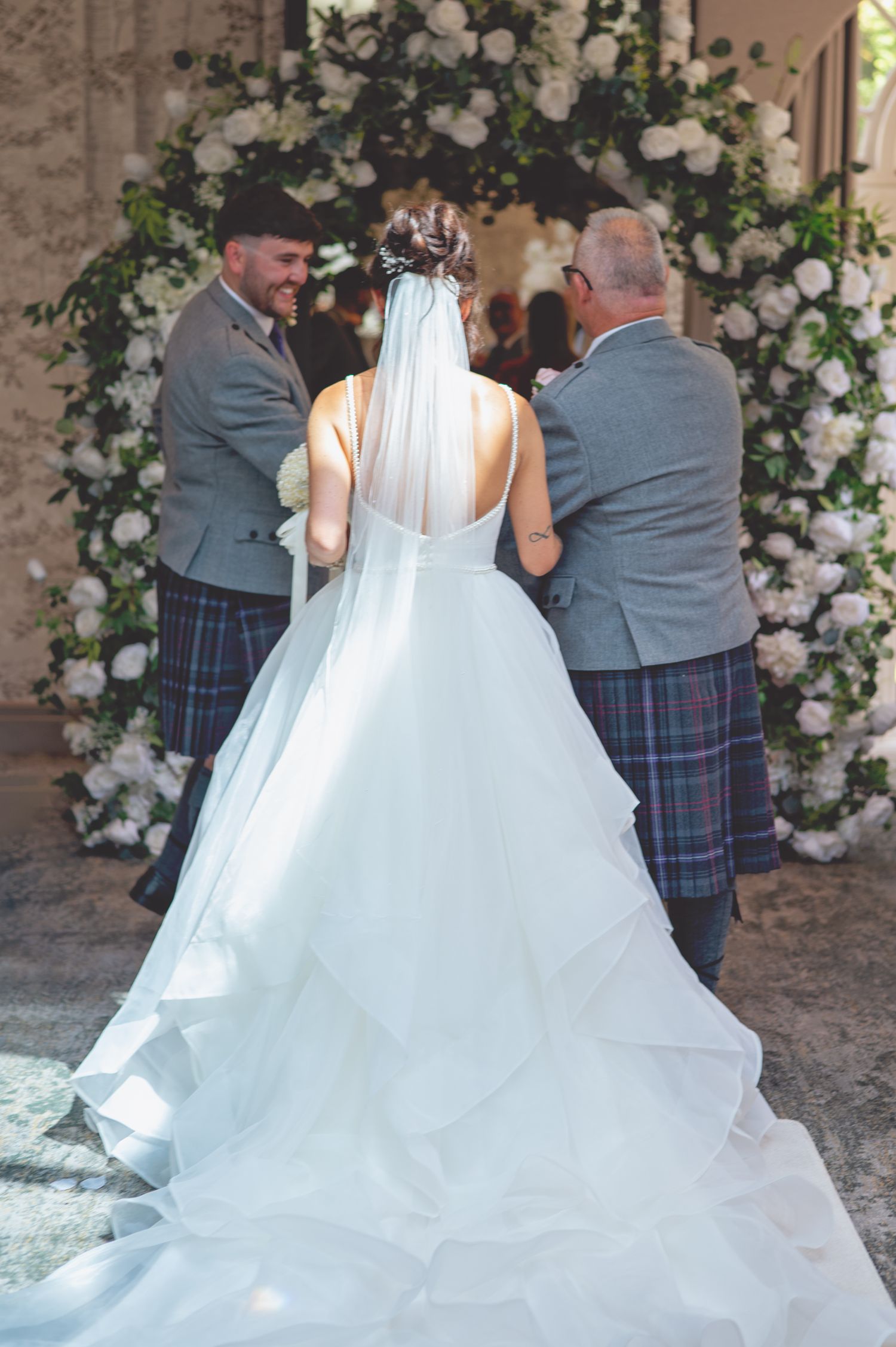A bride in a flowing white gown stands before a floral arch with attendants wearing traditional Scottish kilts.