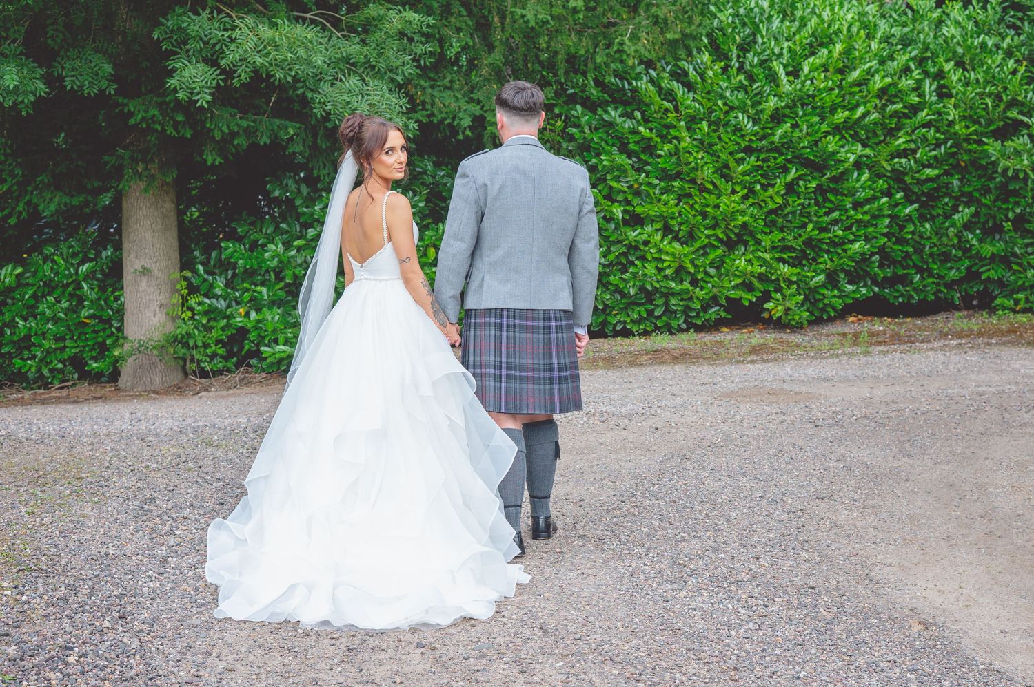 Bride in flowing white gown and groom in kilt walking away on garden path.