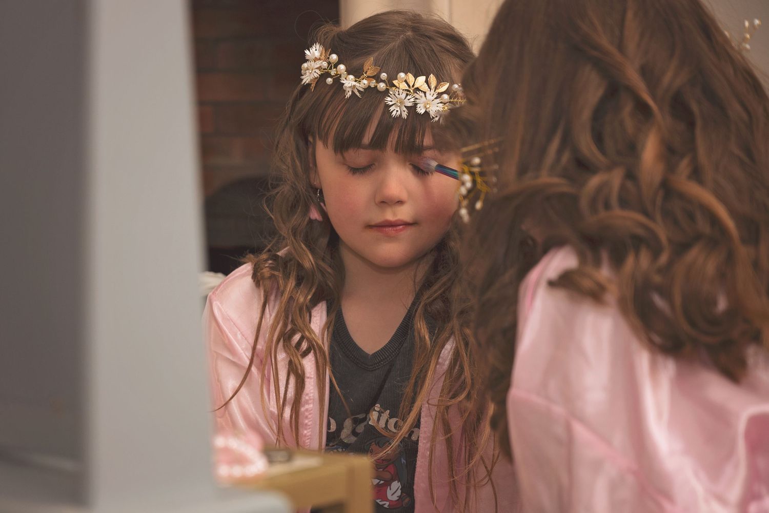 Young girl with floral crown and long brown hair in pink jacket looks down thoughtfully at a wedding event.