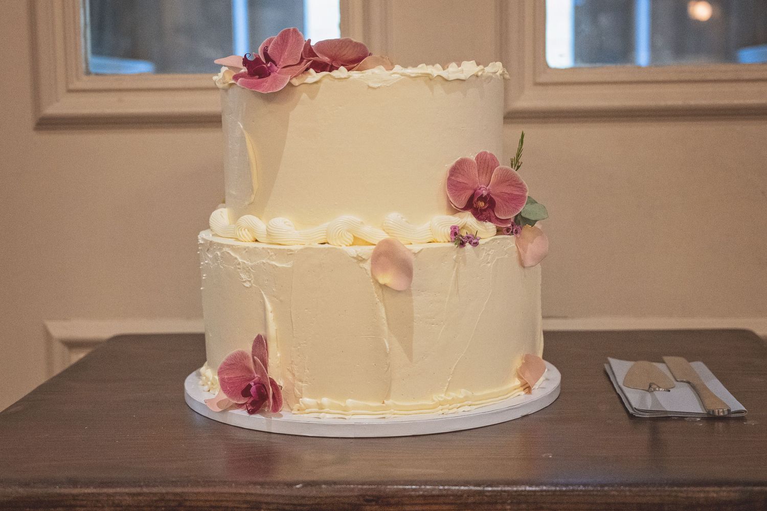 Elegant two-tier white wedding cake decorated with pink orchids and flowers on a wooden table.