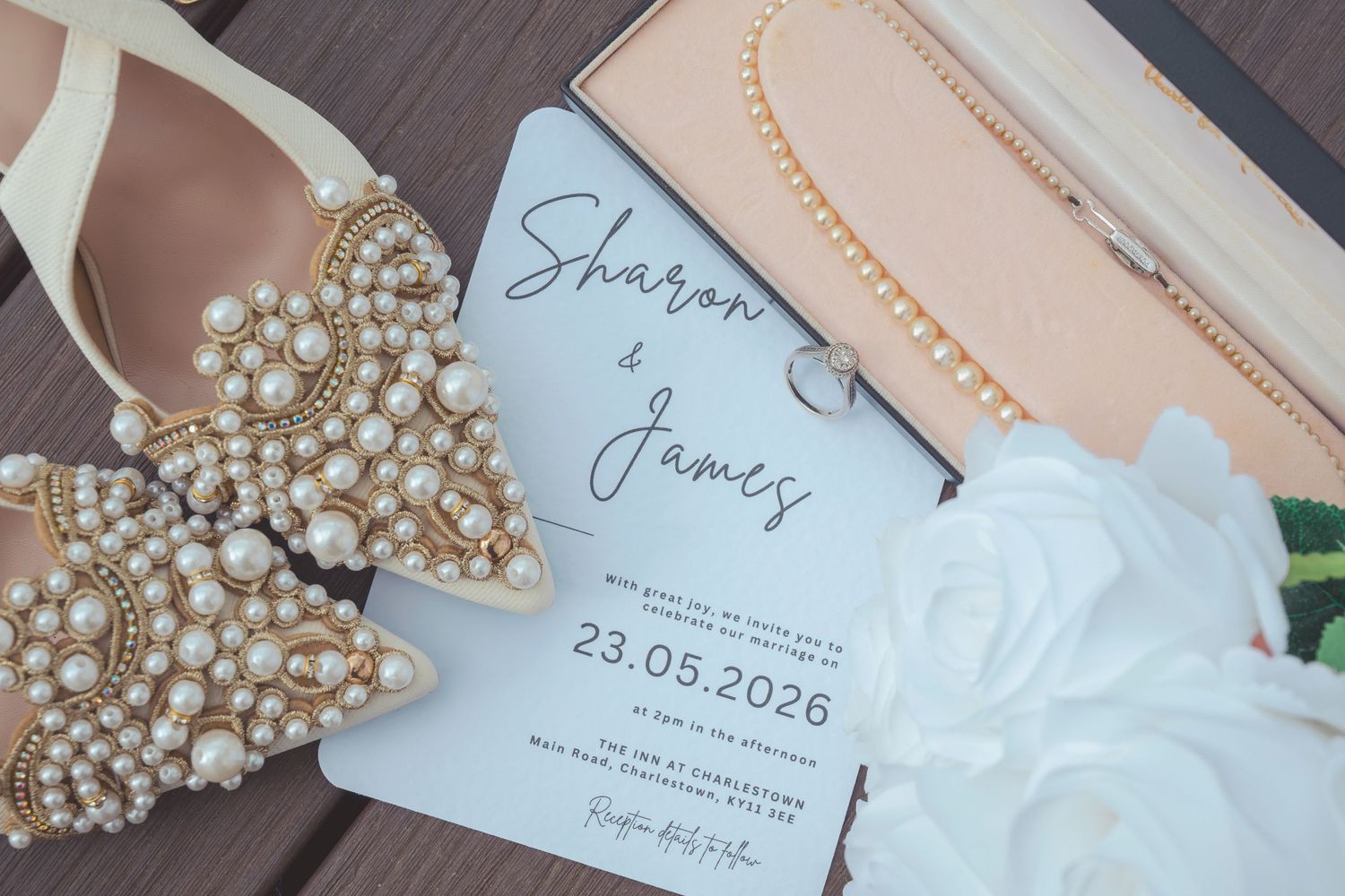 Wedding flat lay with pearl-embellished heels, save the date card for Sharon & James 23.05.2026, pearl necklace, and white roses.