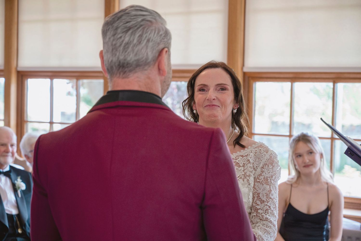 Bride and groom exchanging vows at wedding ceremony indoors, with guests watching in background.