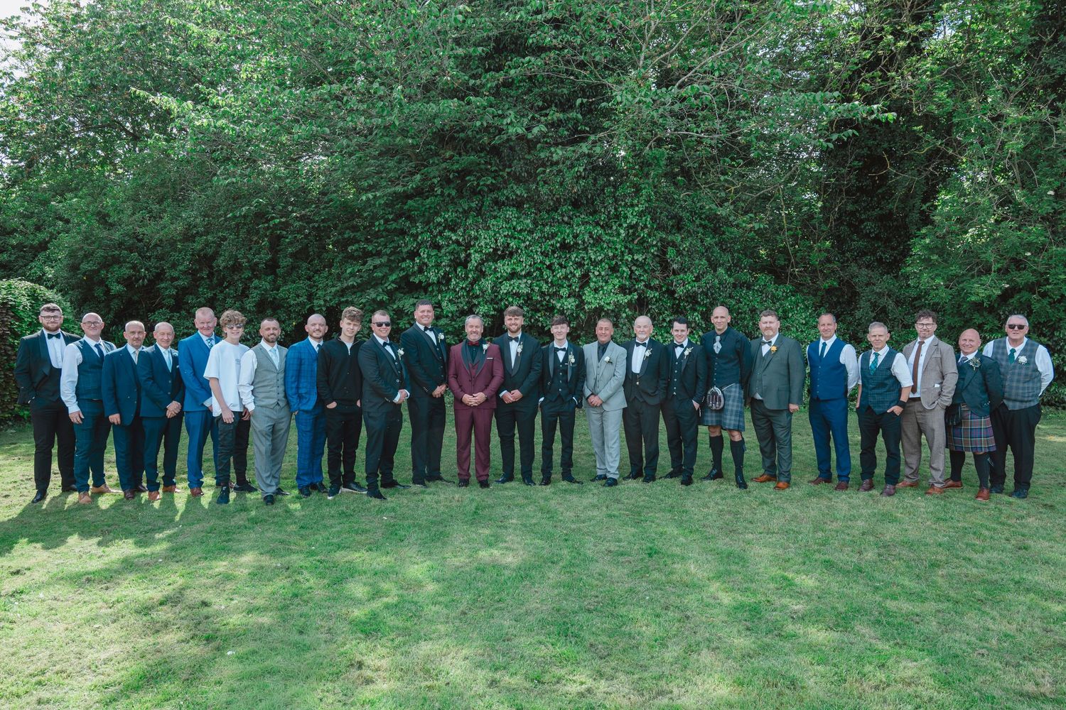 Large group of men in formal attire posing outdoors on green lawn with trees at a wedding celebration.