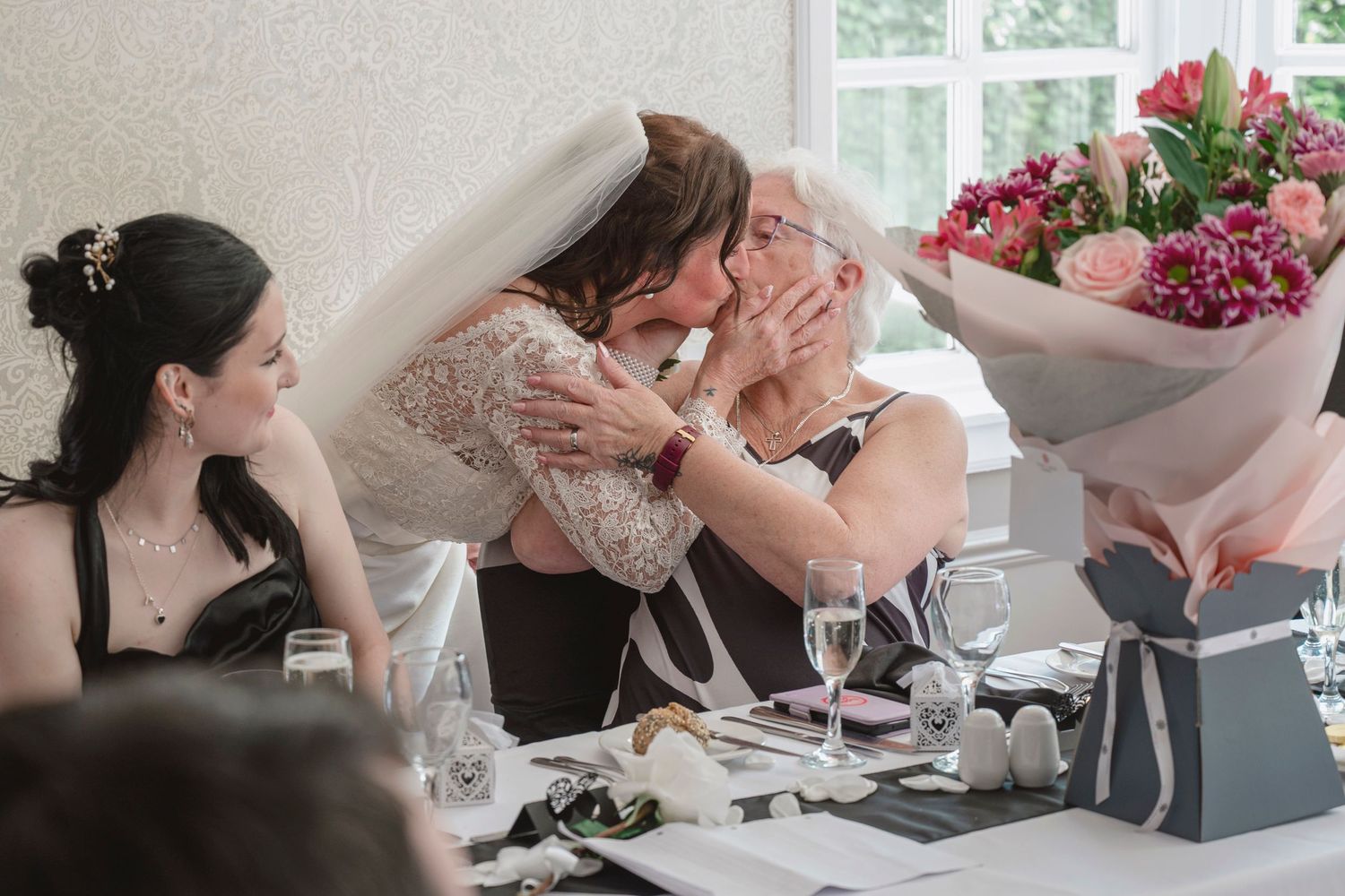 Bride kissing elderly woman at wedding reception table with bridesmaid and pink flower bouquet nearby.