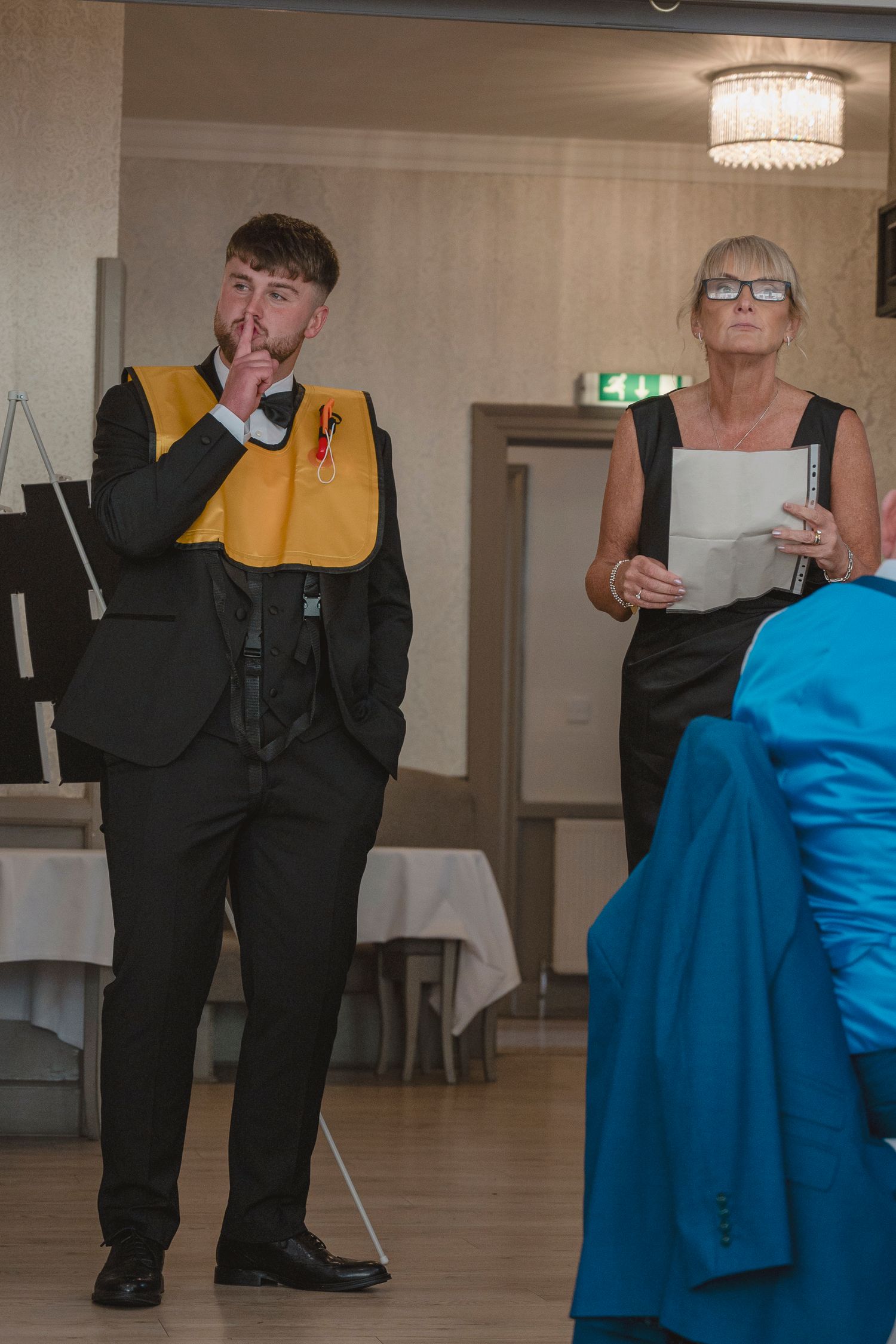 Young man in black suit with yellow sash stands beside older woman holding papers at formal event.