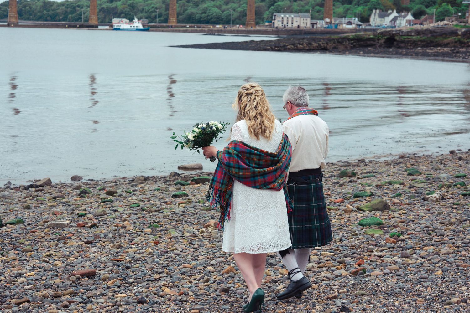 Bride and groom walk along rocky Scottish shoreline, man wearing traditional tartan kilt, woman holding white bouquet.