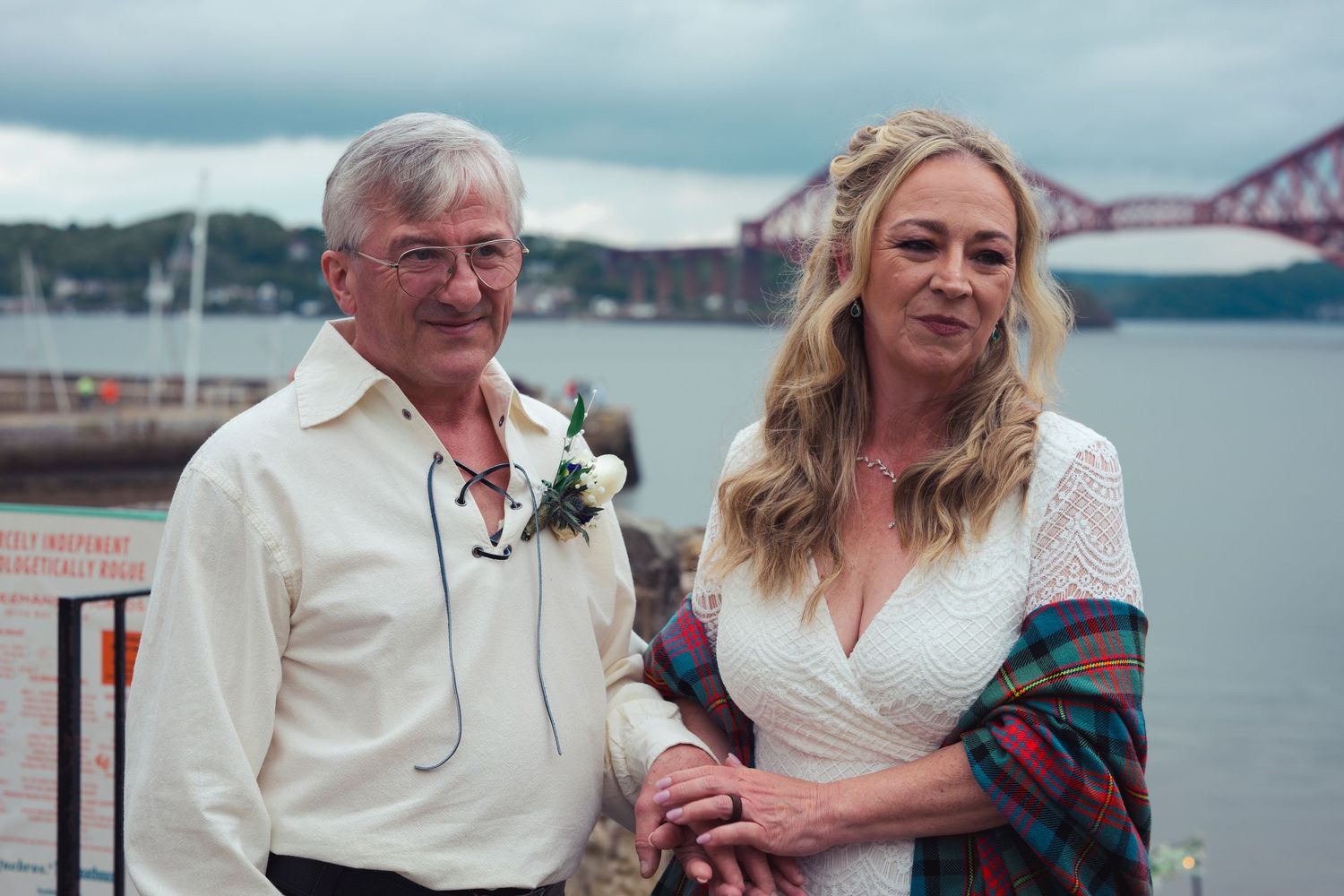 Couple posing by waterfront with Forth Bridge in background, man in white shirt with boutonniere, woman in white lace dress.