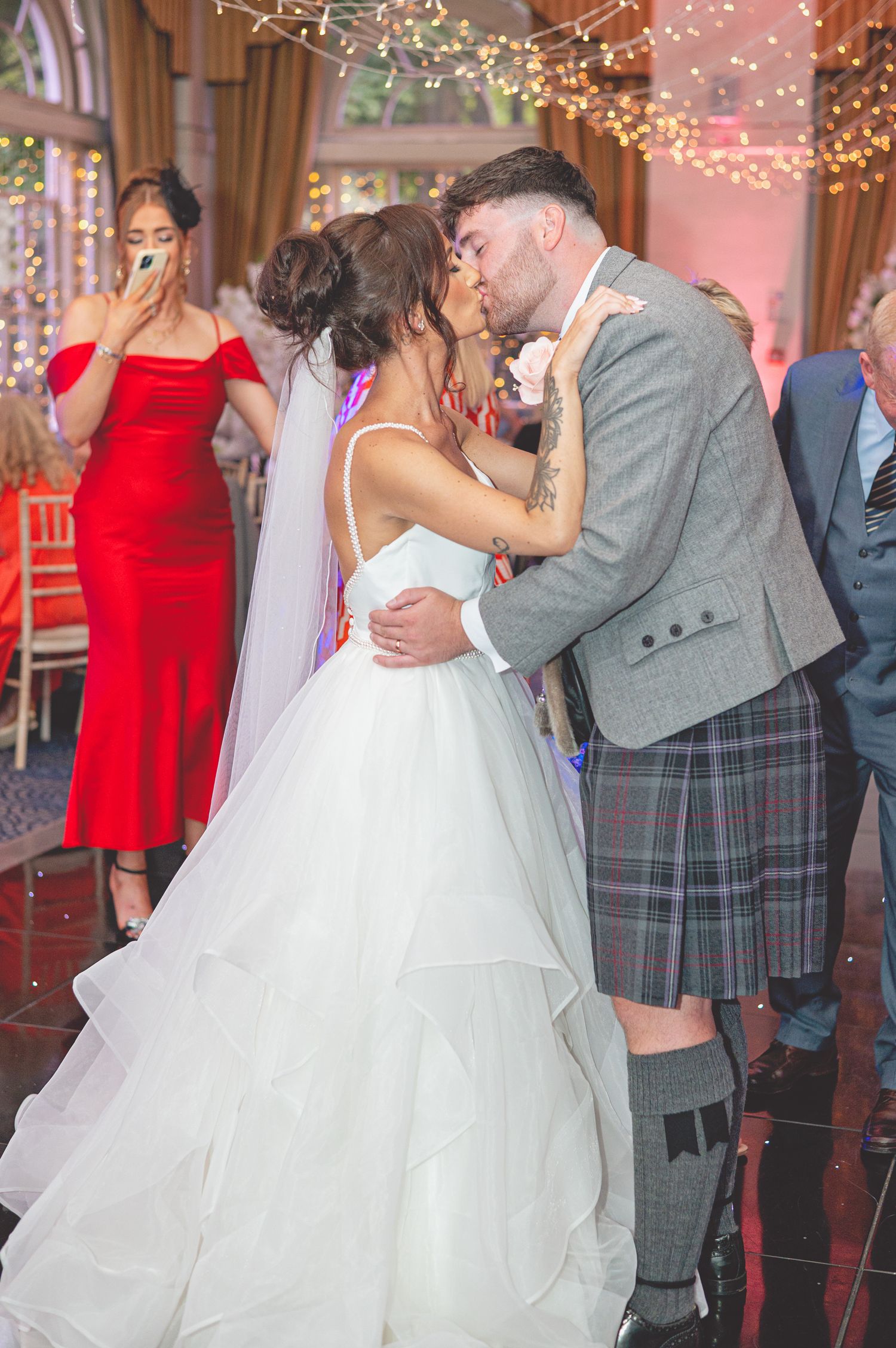 Wedding couple shares a kiss during reception while guest in red dress reacts in background.
