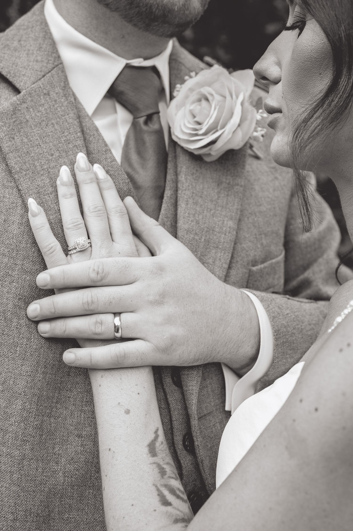 Close-up black and white photo showing wedding rings and a white rose boutonniere on a gray suit.