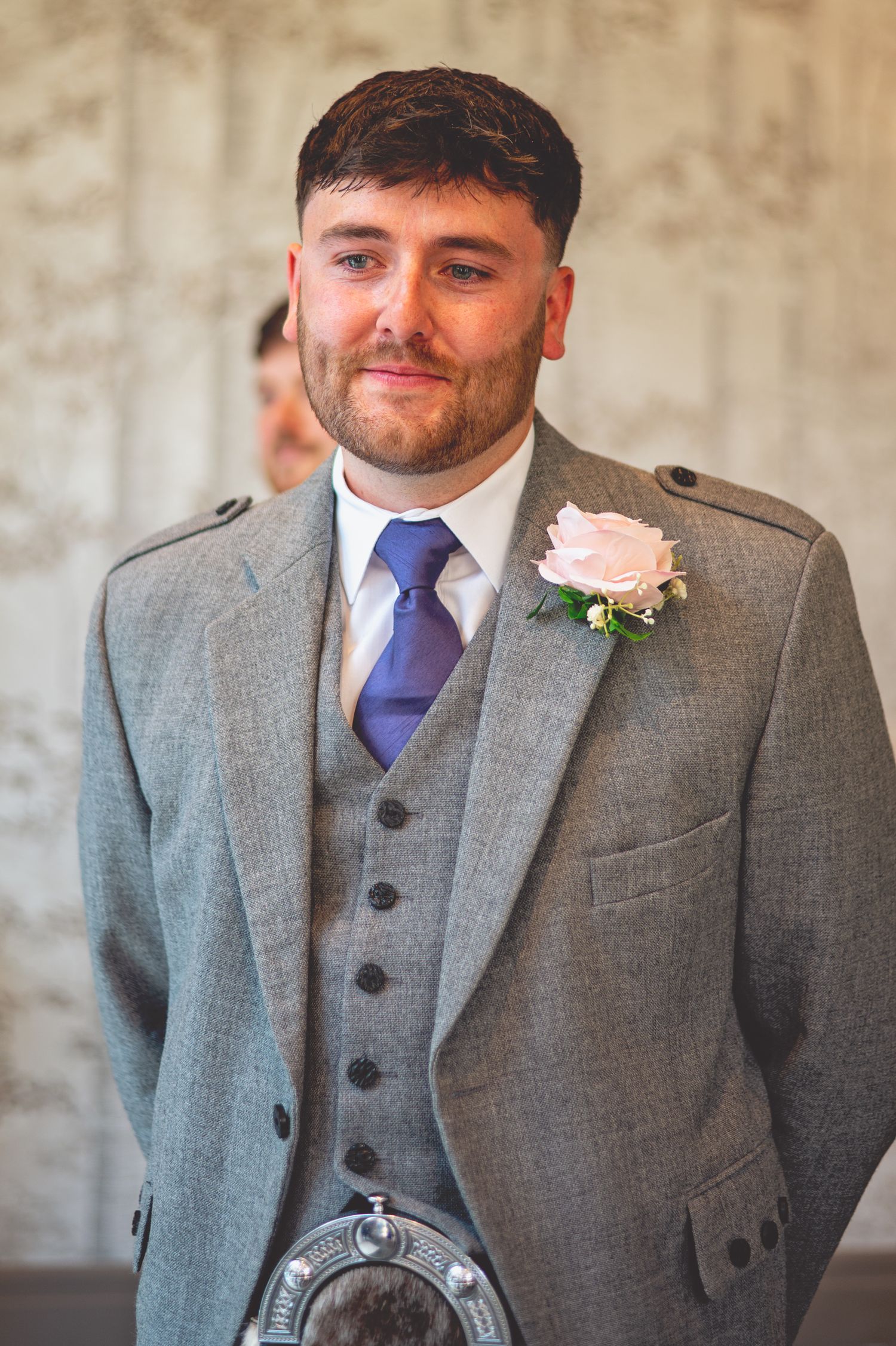Groom in grey three-piece suit with purple tie and Scottish sporran smiling.