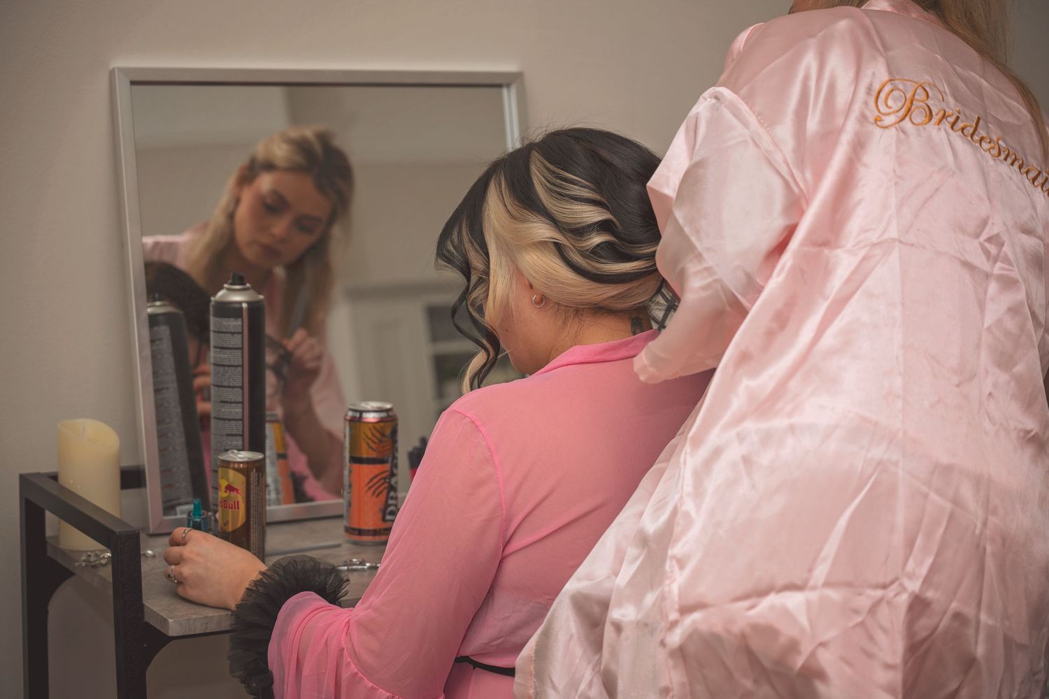 Bridesmaid in pink robe having hair styled with curling iron, reflected in mirror during wedding preparations.