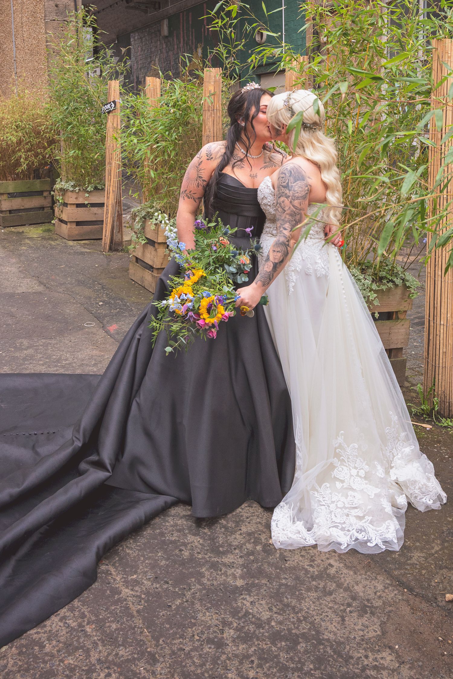 Two tattooed brides kiss outdoors, one in a black ballgown and one in white lace, holding sunflower bouquet.