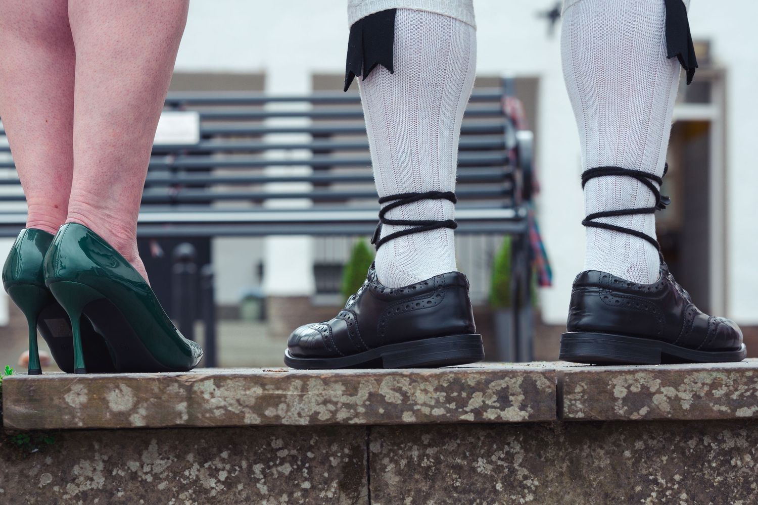 Close-up of two people standing on a stone wall, one wearing green heels and the other black brogues with white socks.