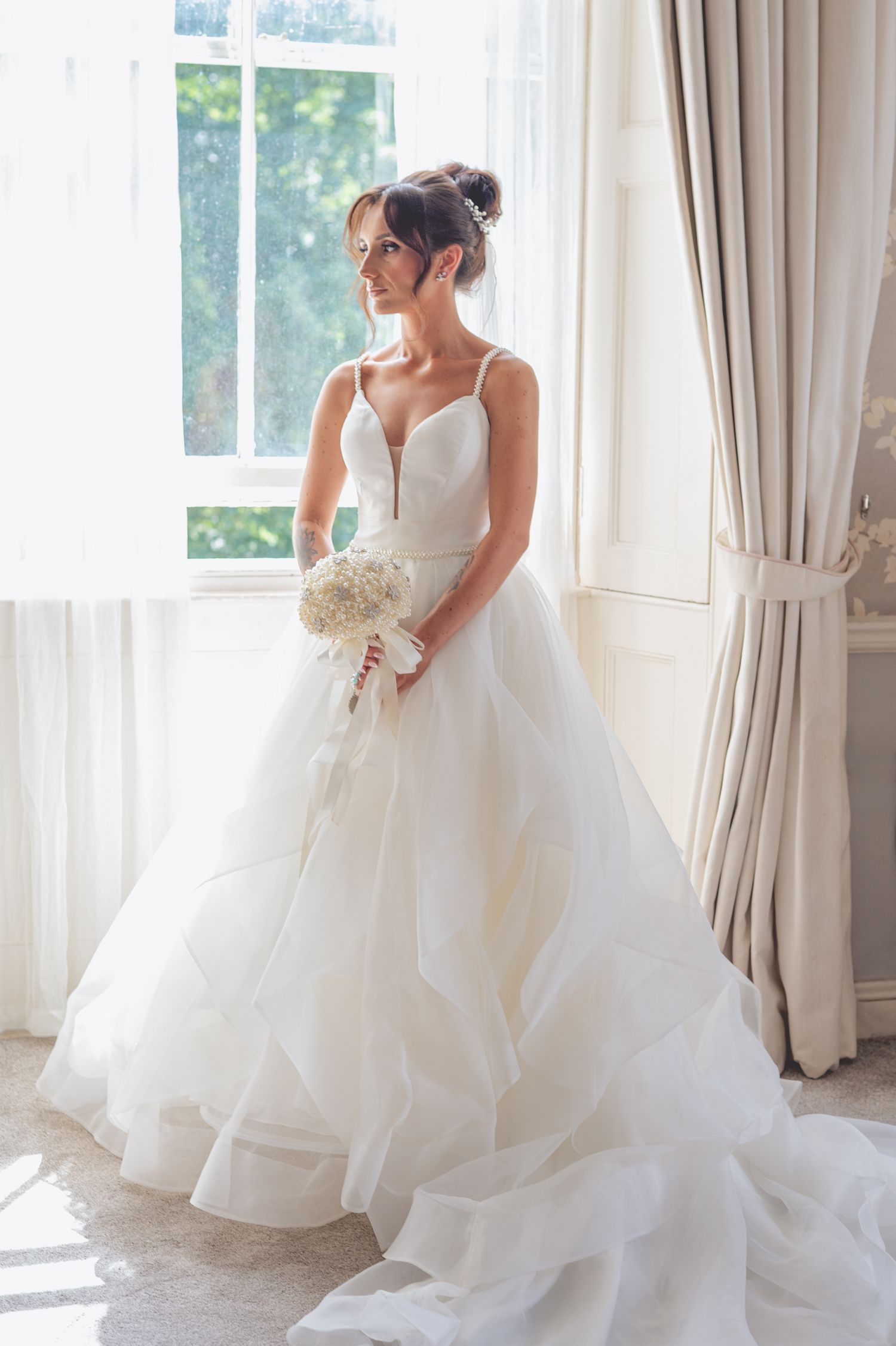 Bride in flowing white ballgown poses elegantly by sunlit windows.