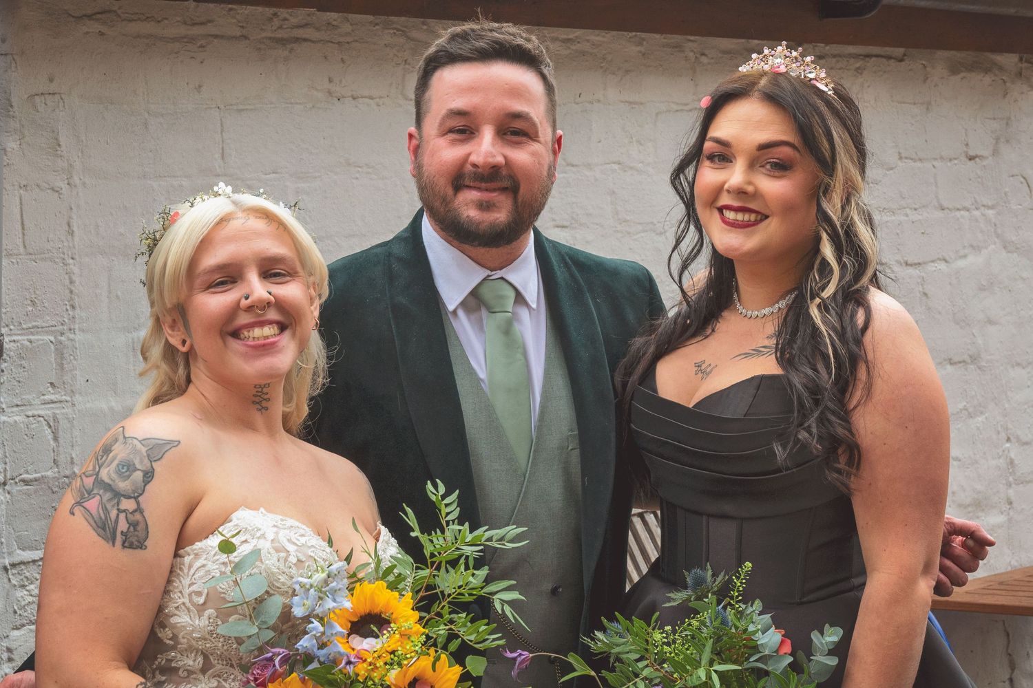 Two brides in wedding attire smile with groom in green suit holding colorful floral bouquets after ceremony.