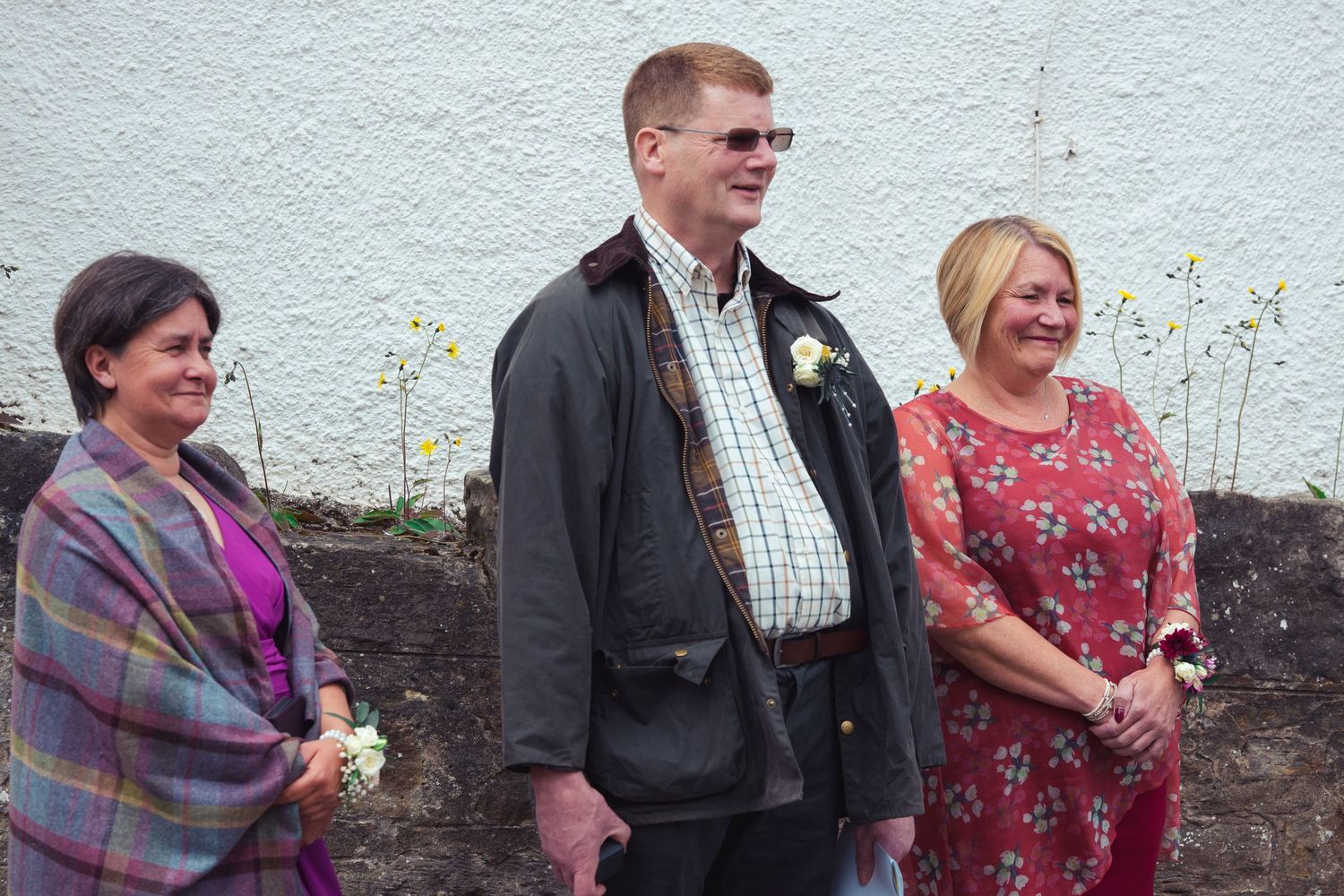 Three smiling adults standing outdoors against a white wall, dressed in smart casual attire at a celebratory event.