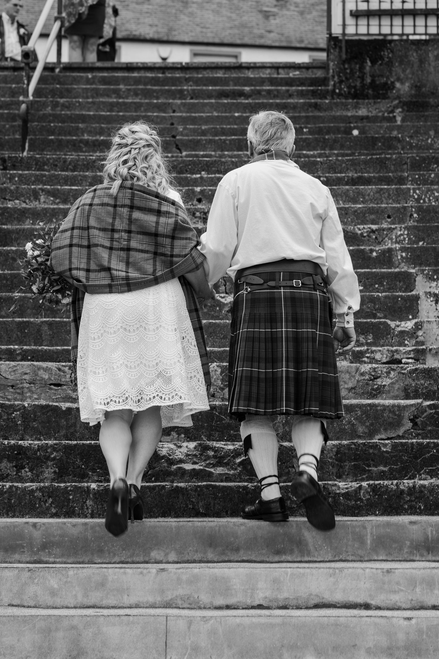 Black and white photo of a couple in Scottish attire climbing stone steps, bride holding flowers, groom wearing a kilt.