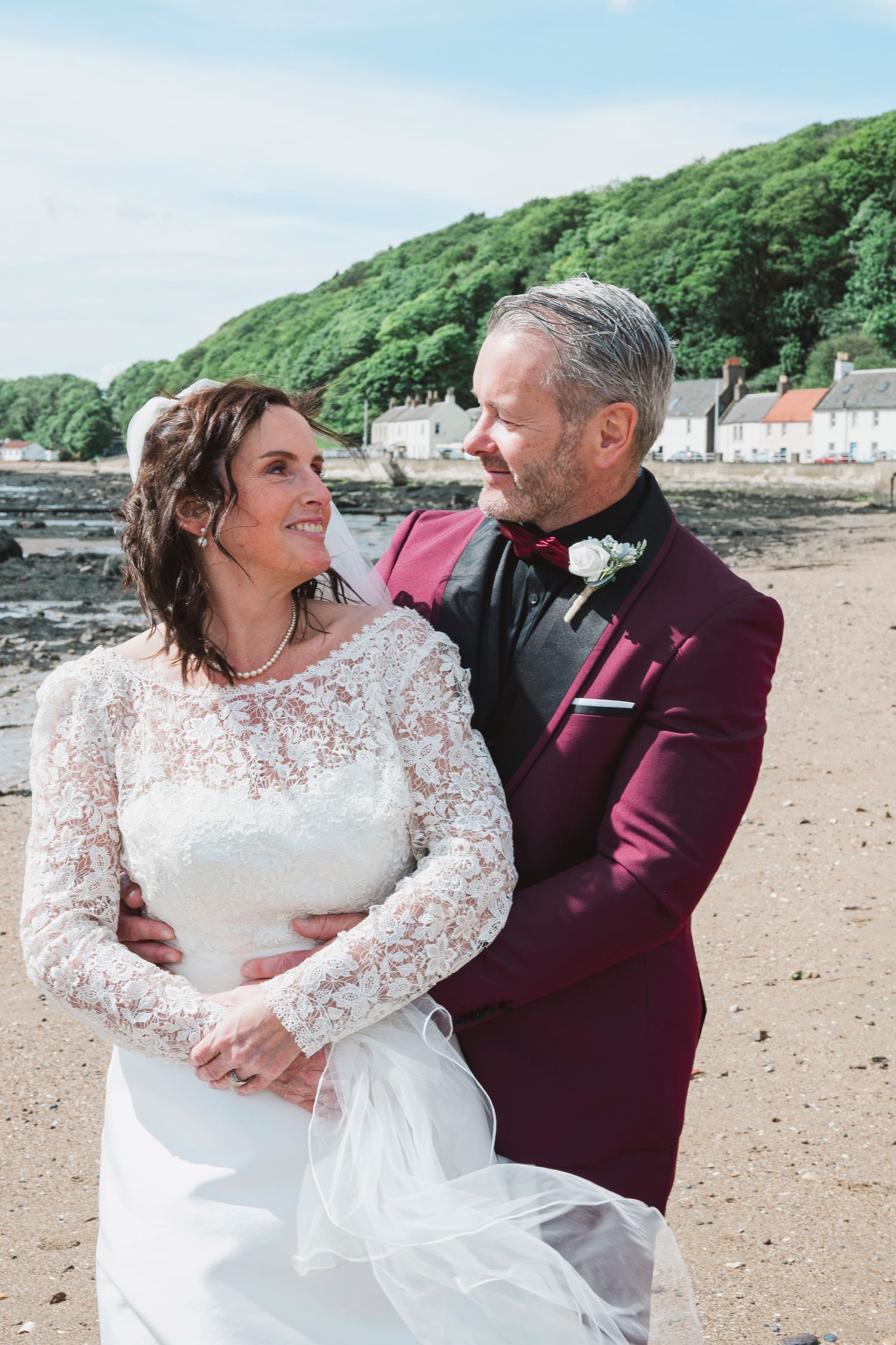 A bride and groom share a romantic moment on a beach, she in lace gown, he in burgundy suit.