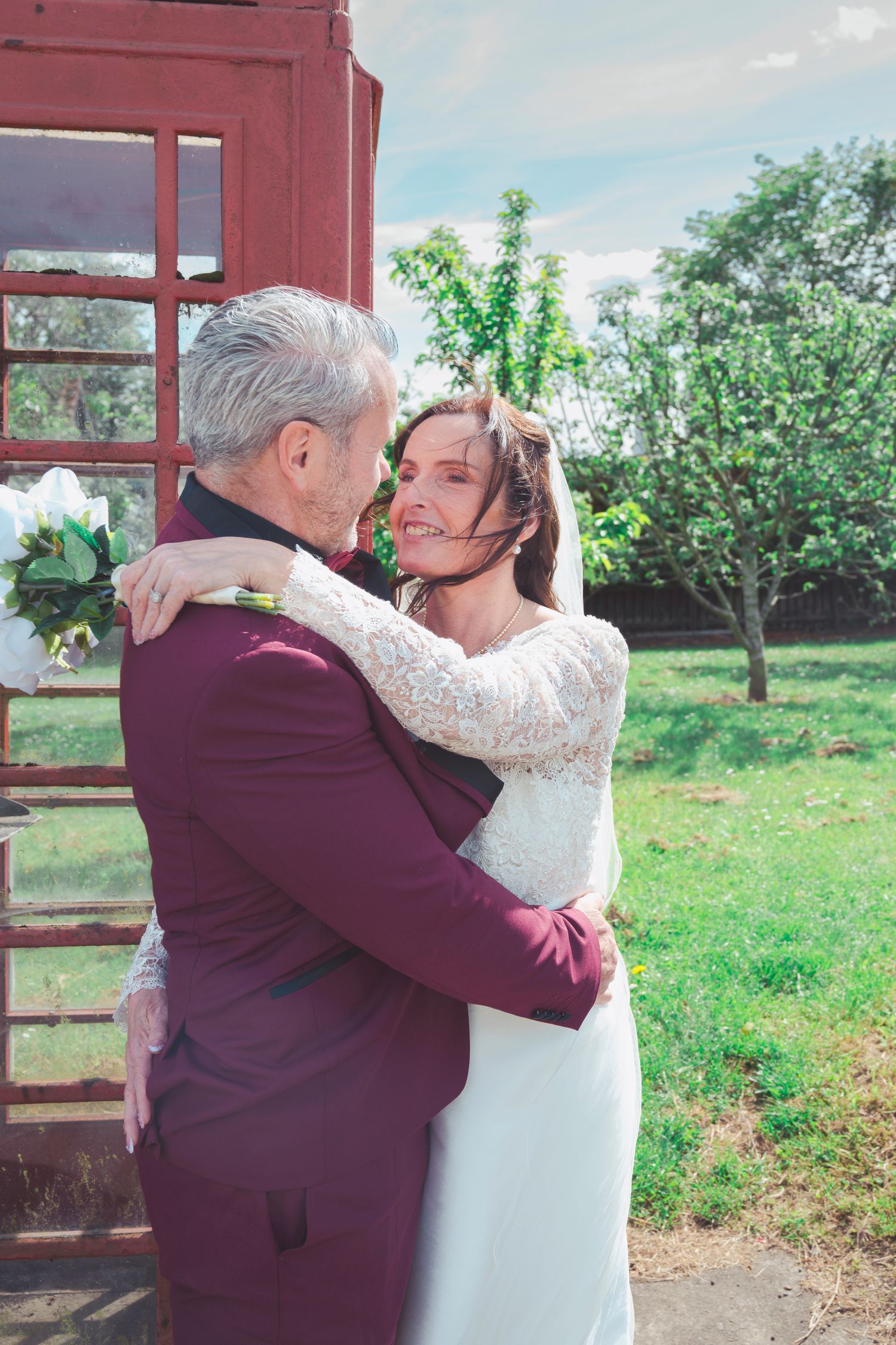 Bride and groom embracing outdoors near red phone booth, wearing white lace gown and burgundy suit on wedding day.