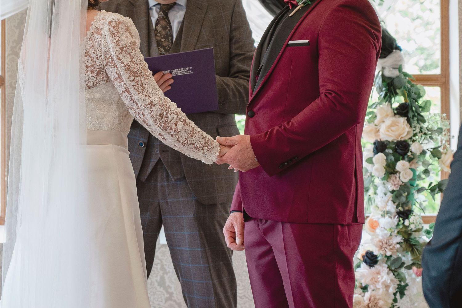 Groom in burgundy suit holds bride's hands at wedding altar with officiant in grey suit and floral backdrop.