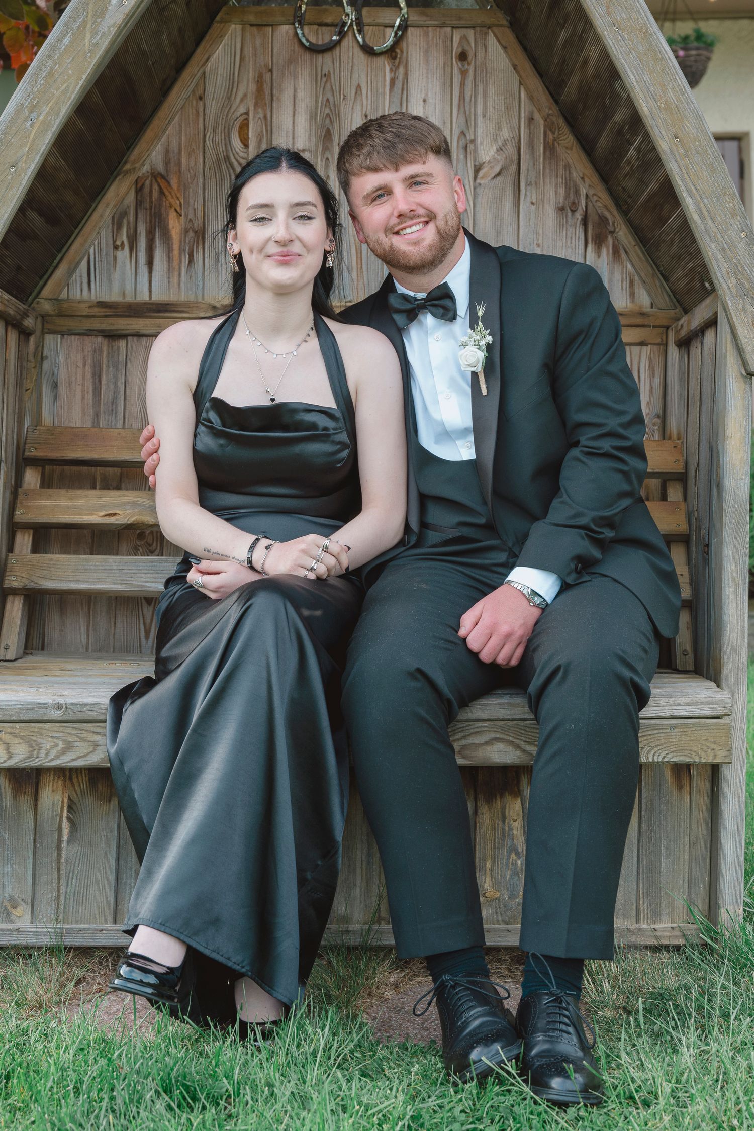 A couple in formal attire sit together in a wooden gazebo, wearing matching black outfits for prom.