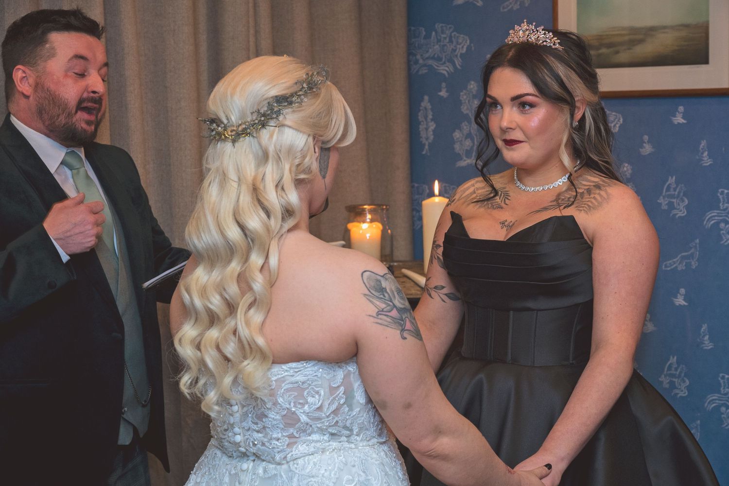 Two brides holding hands during intimate wedding ceremony with officiant and candles in background.