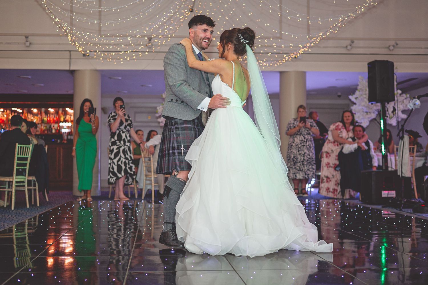 Couple sharing their first dance under twinkling lights on a reflective dance floor.