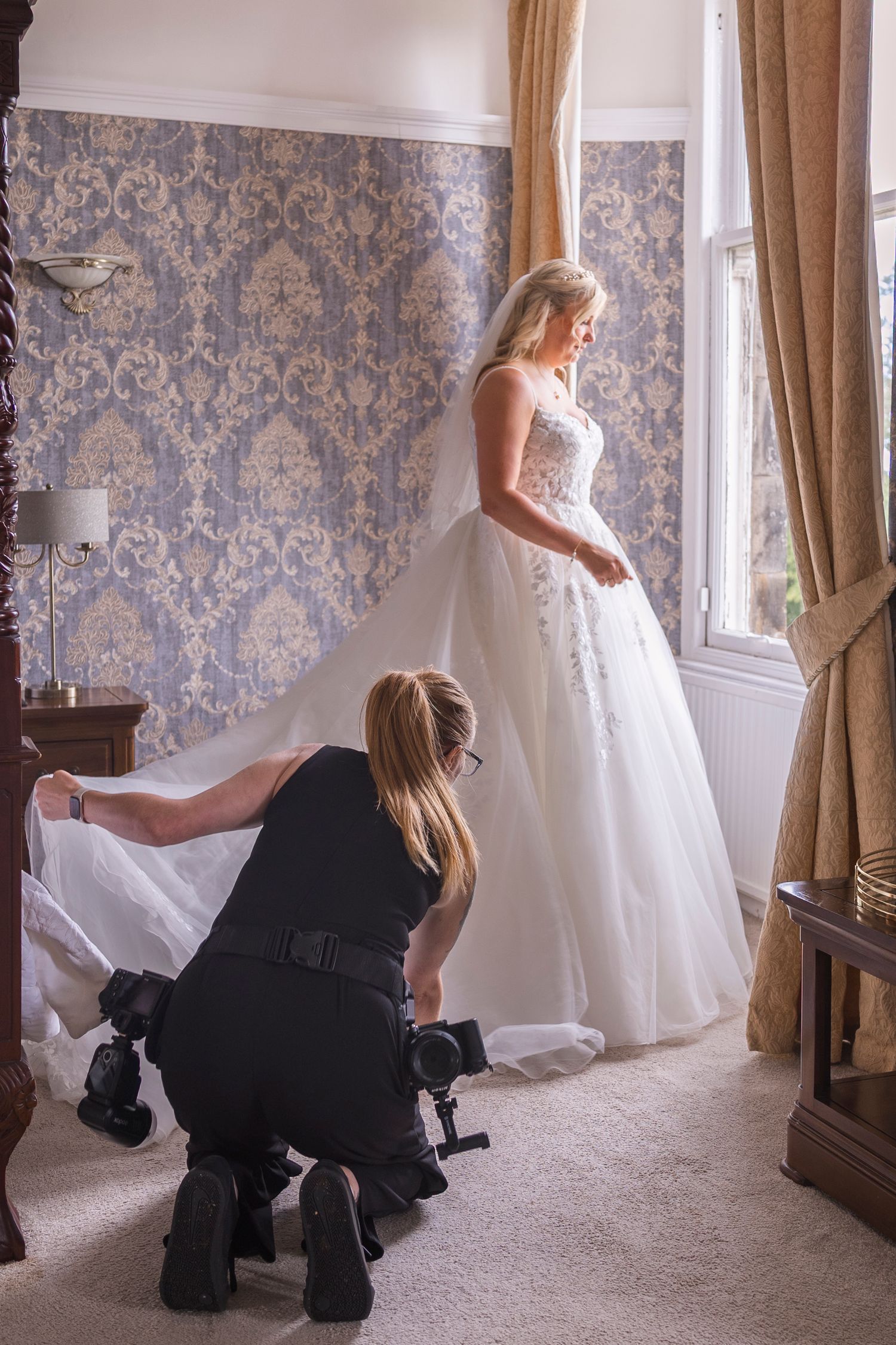 Photographer crouching to arrange wedding dress train of blonde bride standing by window in elegant room.