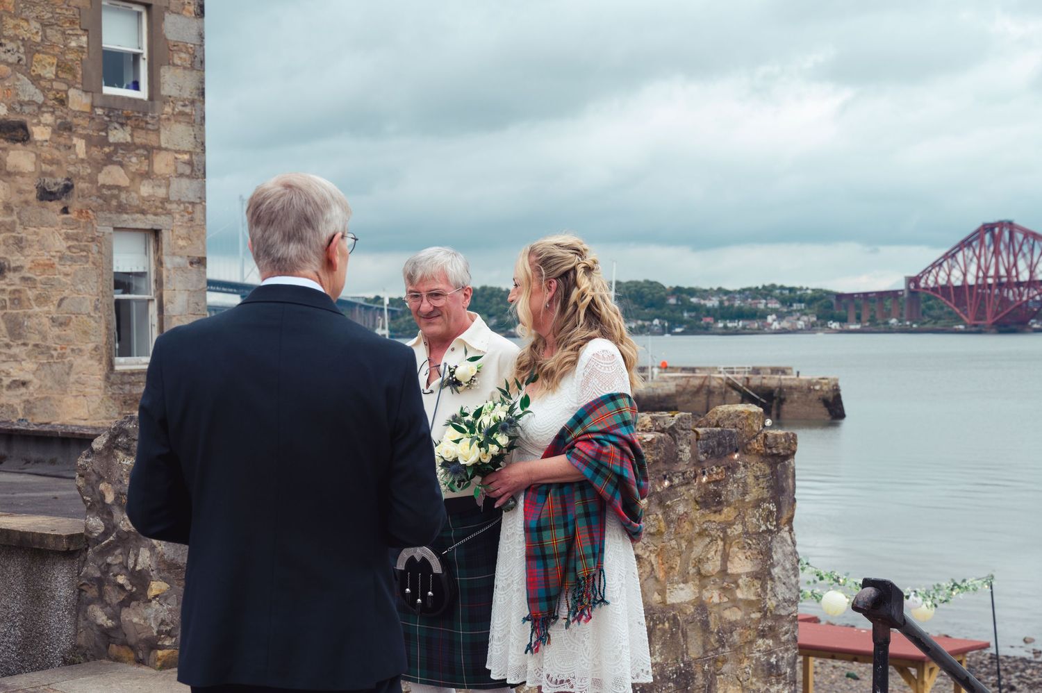 Outdoor wedding ceremony at Scottish castle with bride holding bouquet, groom in kilt, and Forth Bridge visible in background.