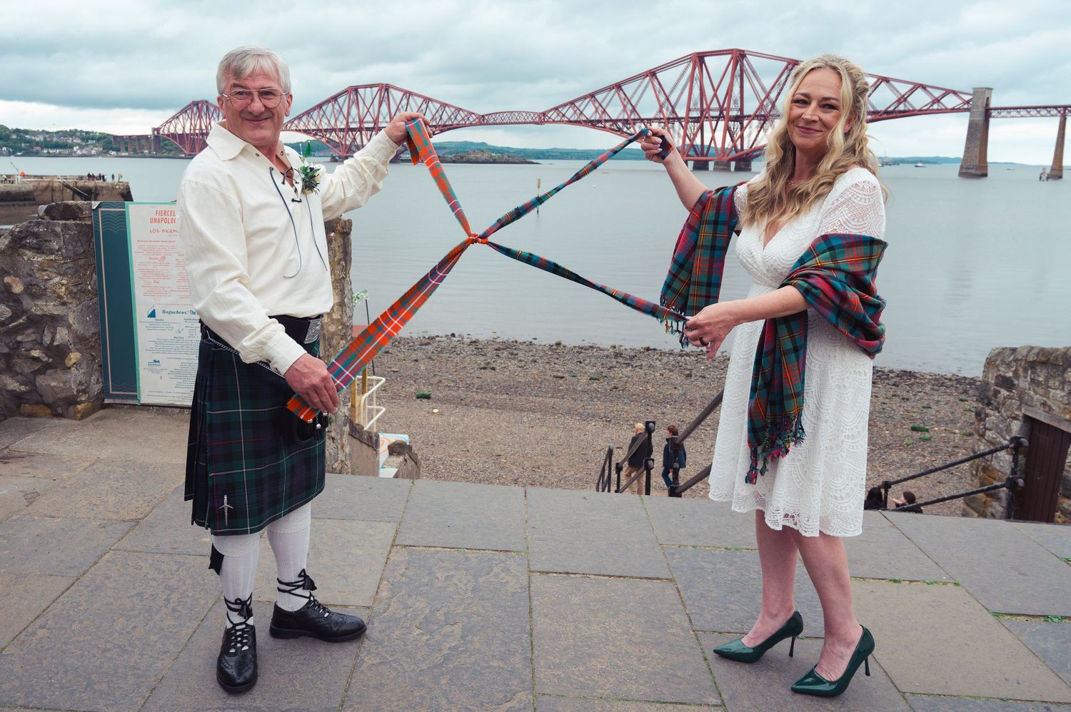 Man in kilt and woman in tartan shawl hold bagpipes near Forth Bridge, Scotland.