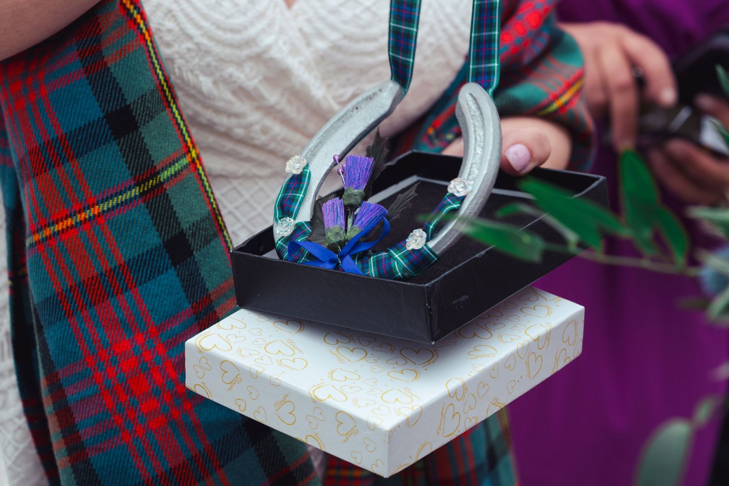 Person in tartan kilt holding a black box with decorative glass flowers and purple blooms at a Scottish ceremony.