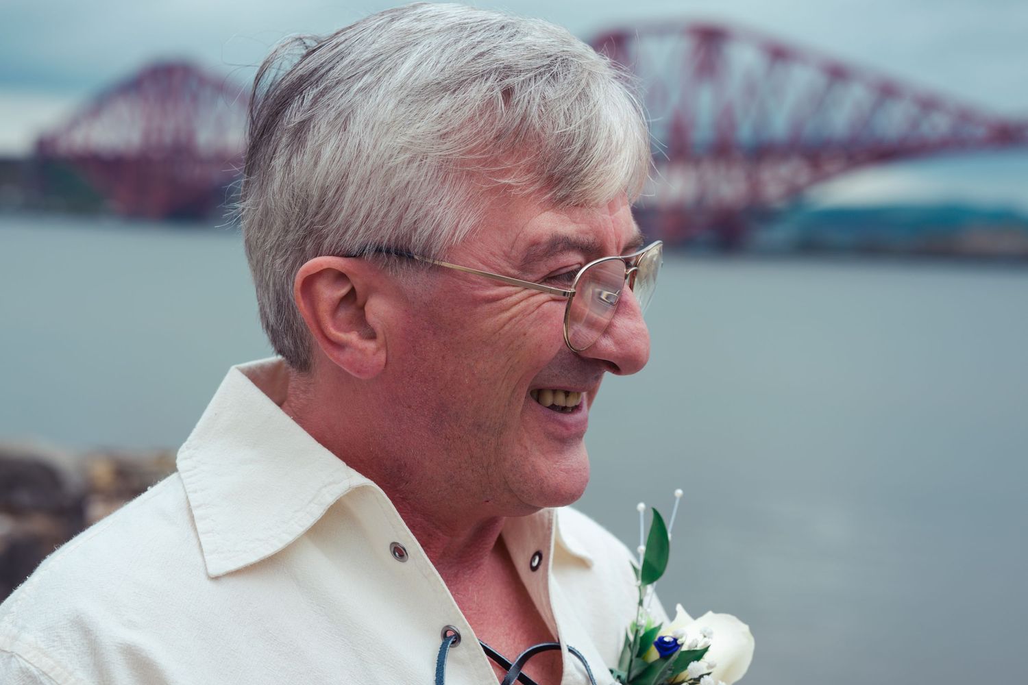 Smiling older man with glasses wearing a boutonniere near the Forth Bridge in Scotland.