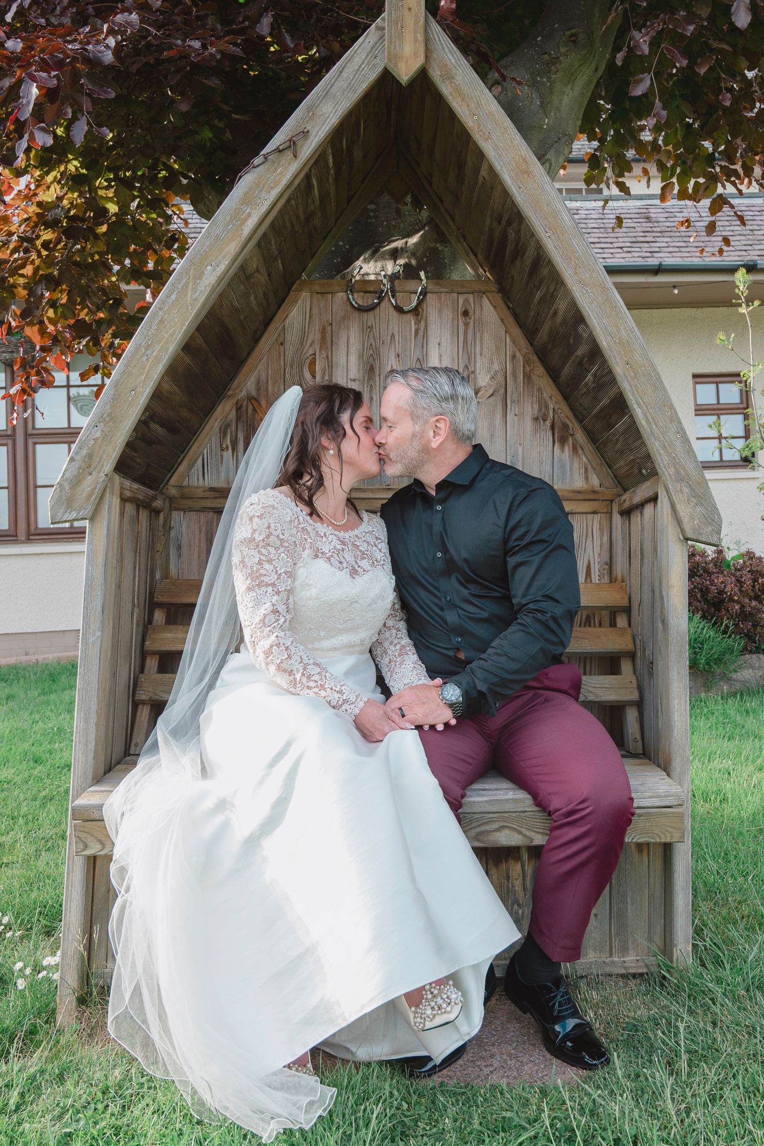Bride and groom share a romantic kiss seated in a rustic wooden arbor bench outdoors on their wedding day.