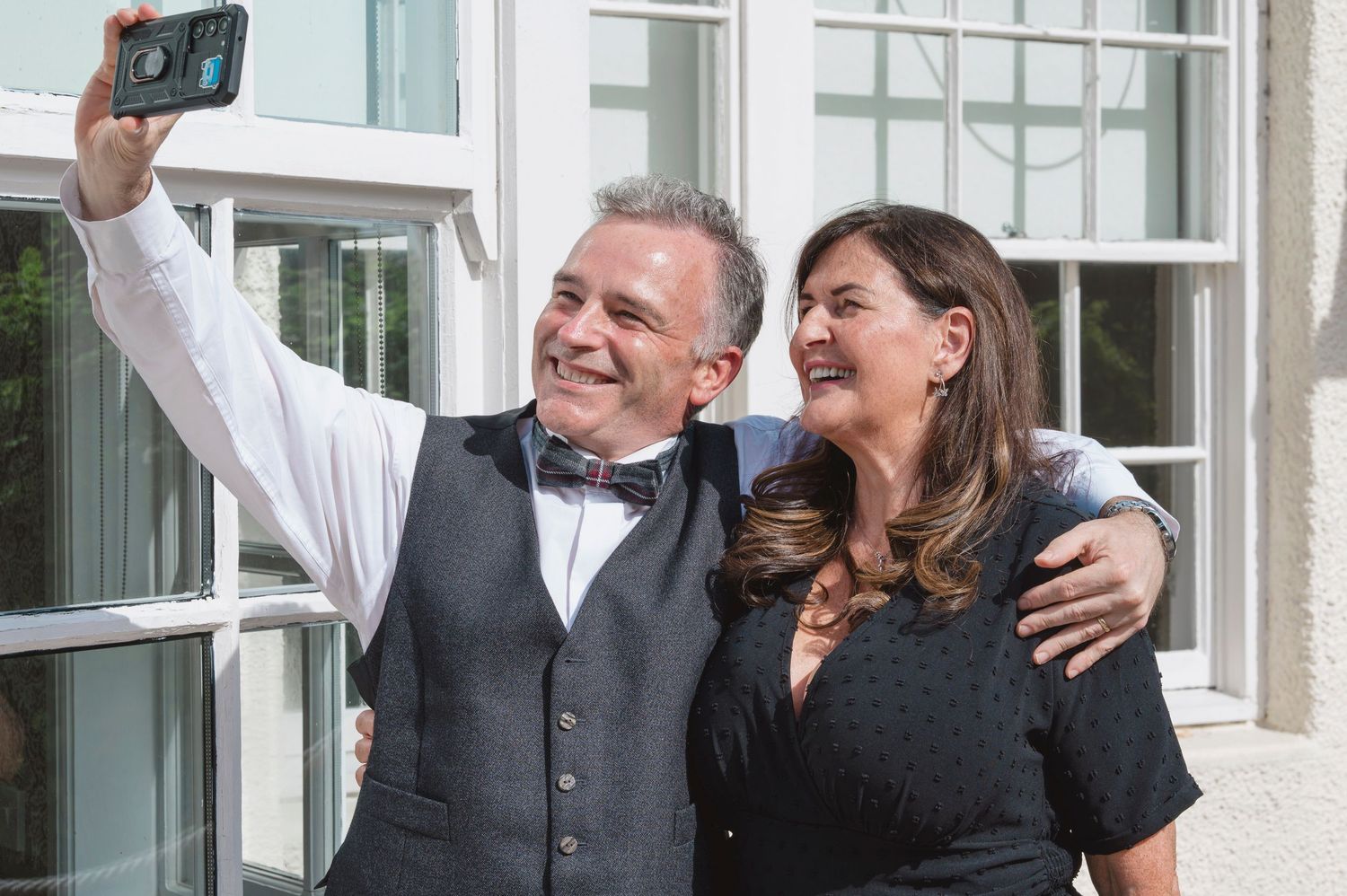 A well-dressed couple smiling and taking a selfie outdoors near a building on a sunny day.