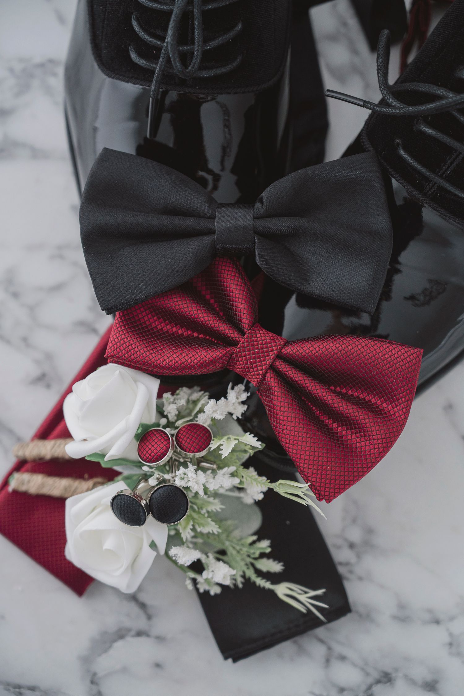 Black patent leather shoes with black and red bow ties, cufflinks, boutonniere, and pocket square on marble surface.