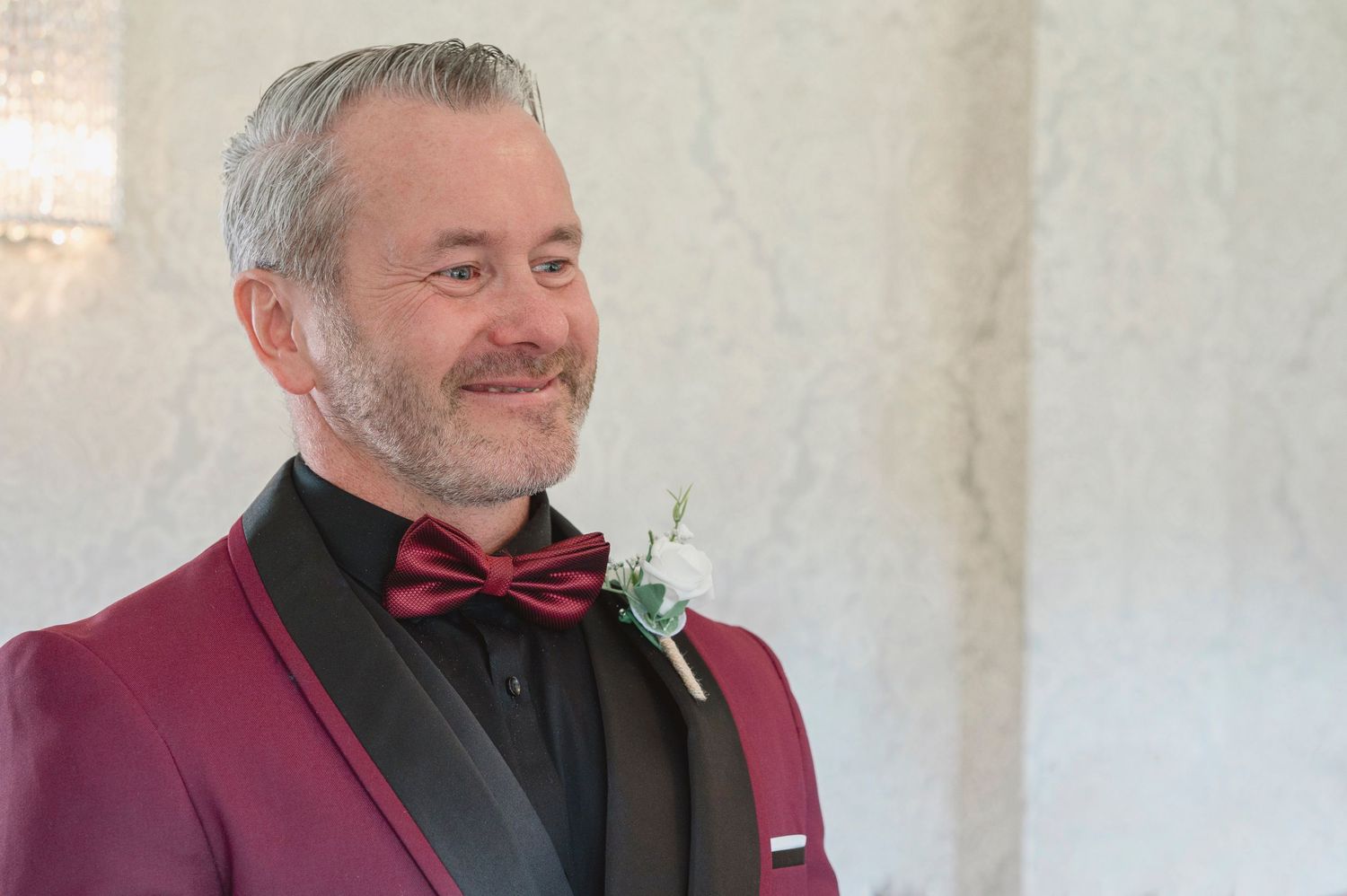 Man in burgundy tuxedo with bow tie and boutonniere smiling at a wedding ceremony.