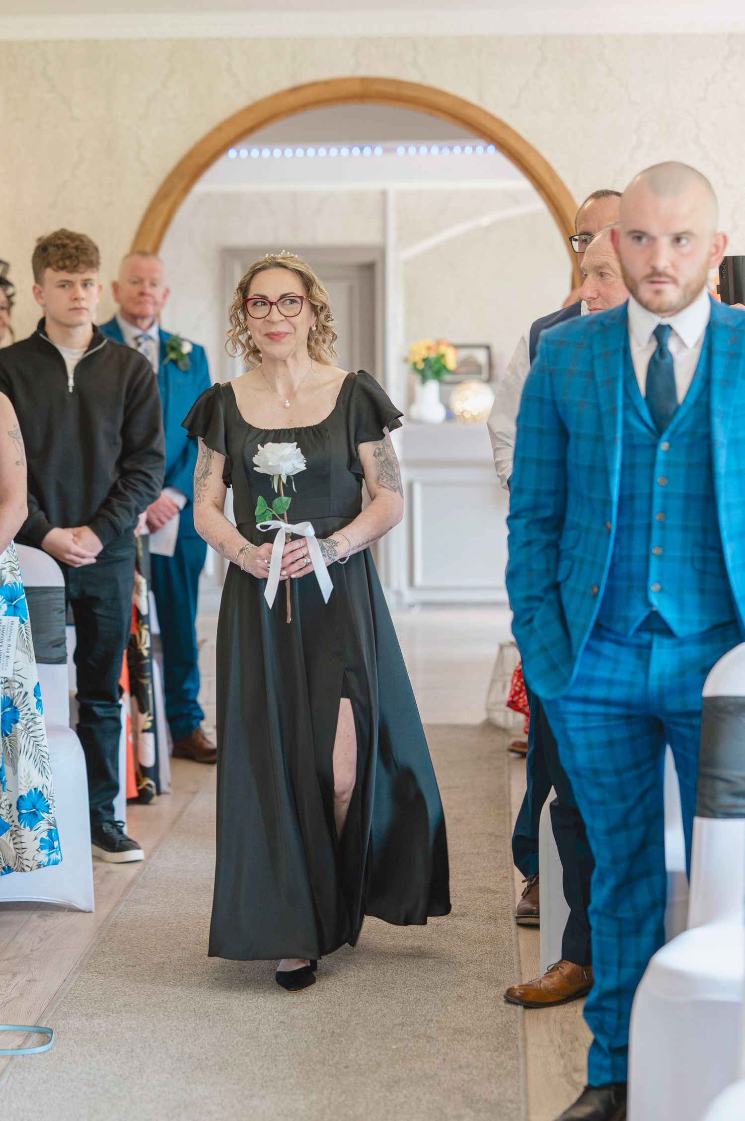 Woman in black dress walks down wedding aisle holding white rose, with guests seated and man in blue suit nearby.