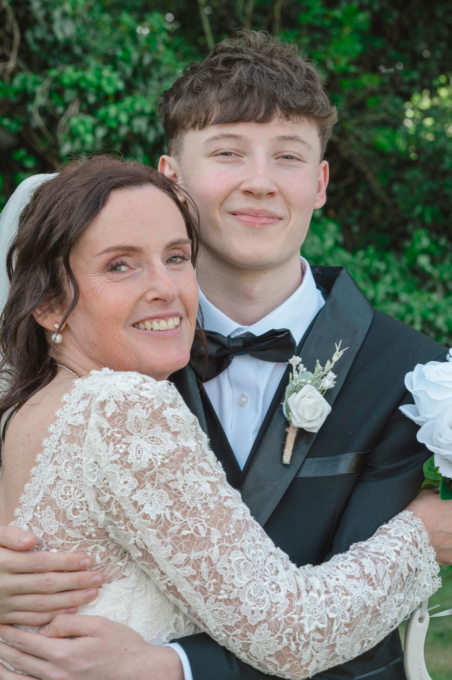 Bride in lace gown embraces young man in black tuxedo with white boutonniere at outdoor wedding ceremony.