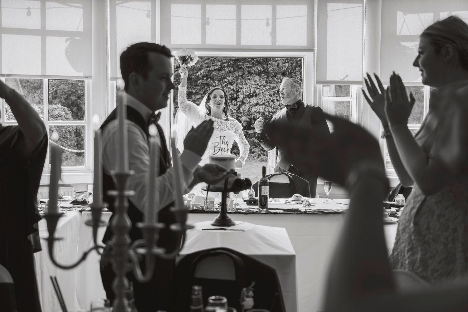Black and white wedding photo of couple cutting cake while guests clap and celebrate in elegant venue.