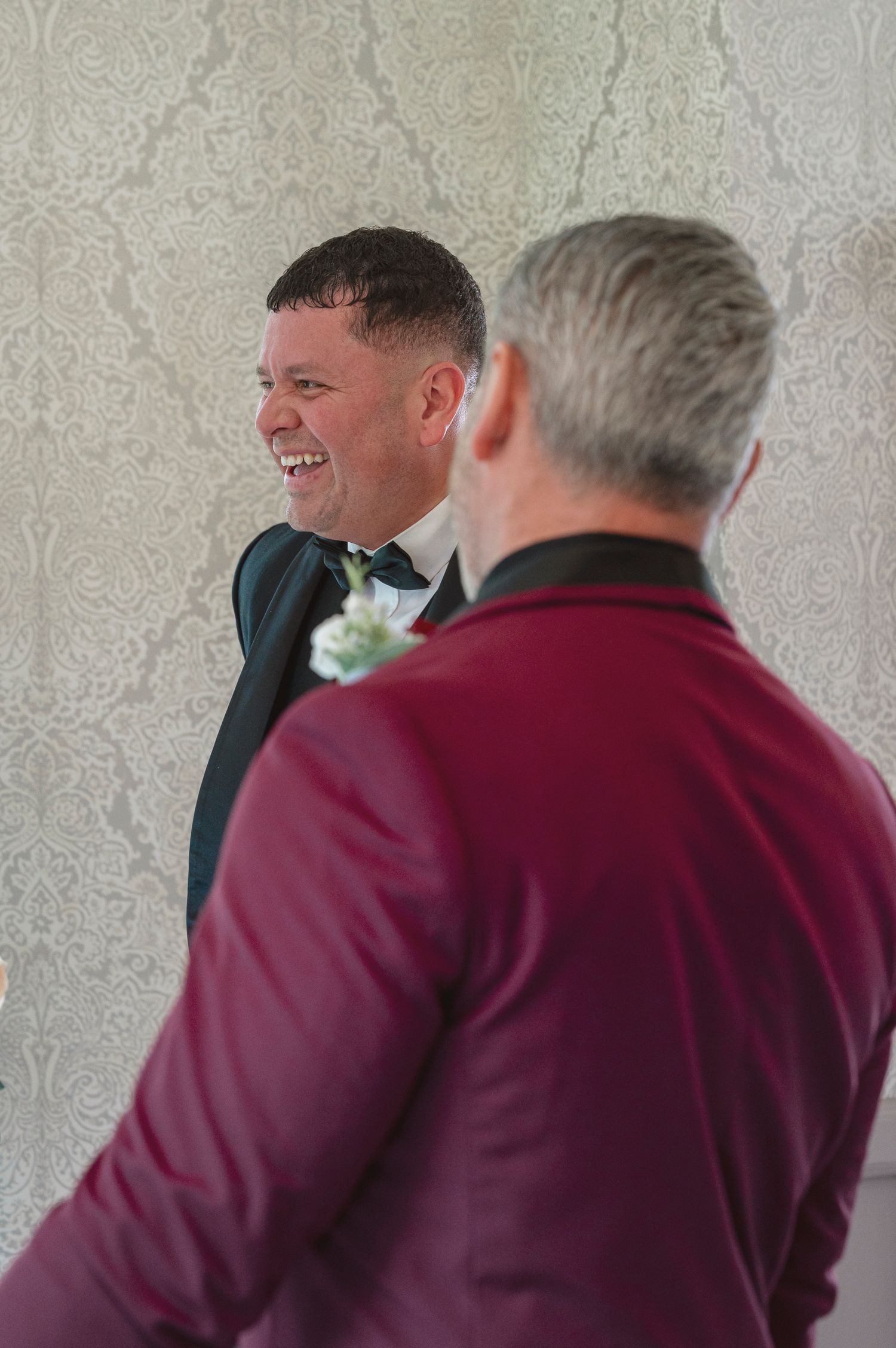 Two men in formal attire share a joyful moment, one wearing a tuxedo with boutonniere, laughing together at a wedding.